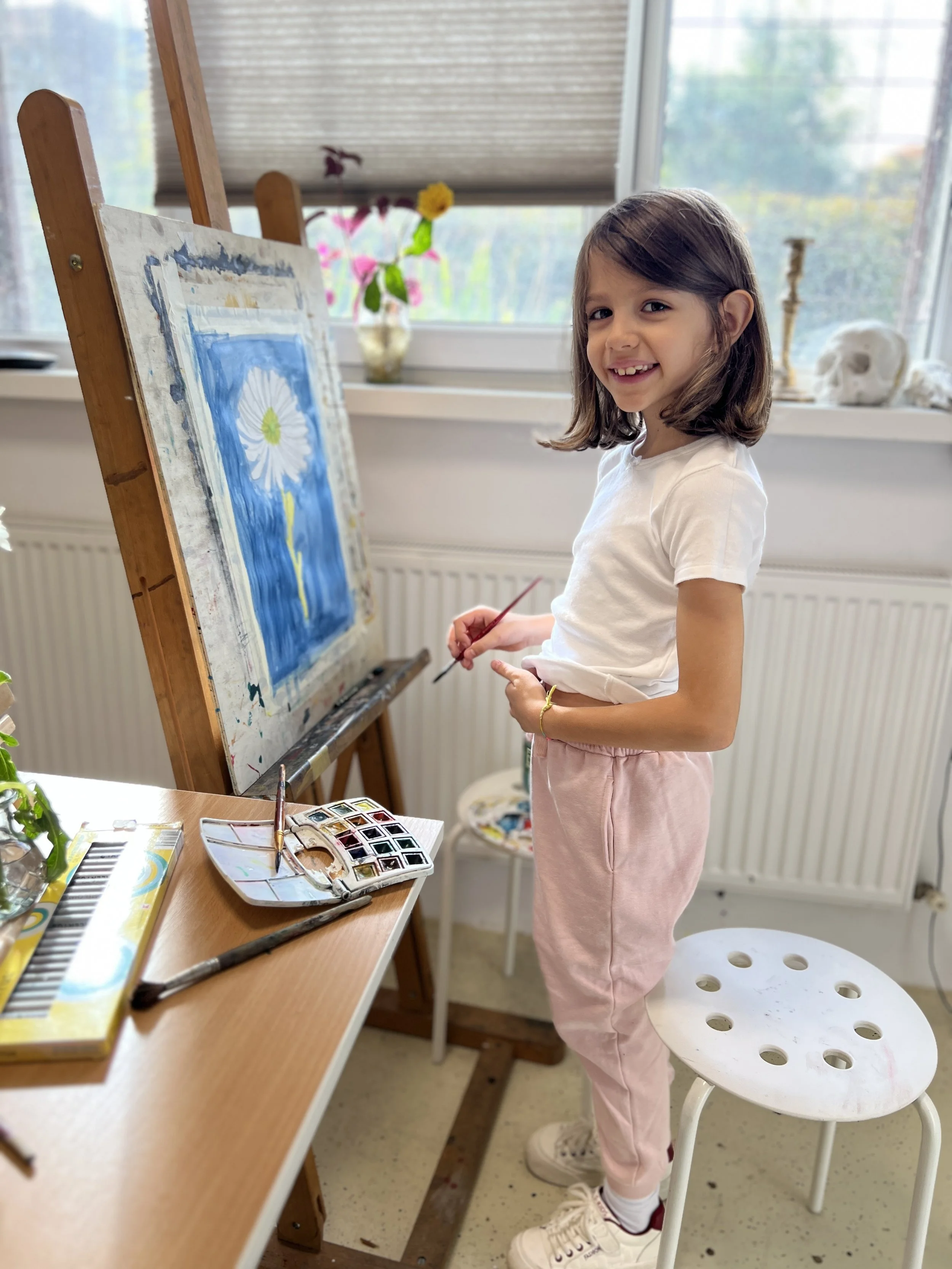 Young girl painting a flower on a canvas in a bright room, with art supplies and window views of plants and skull decorations around her.