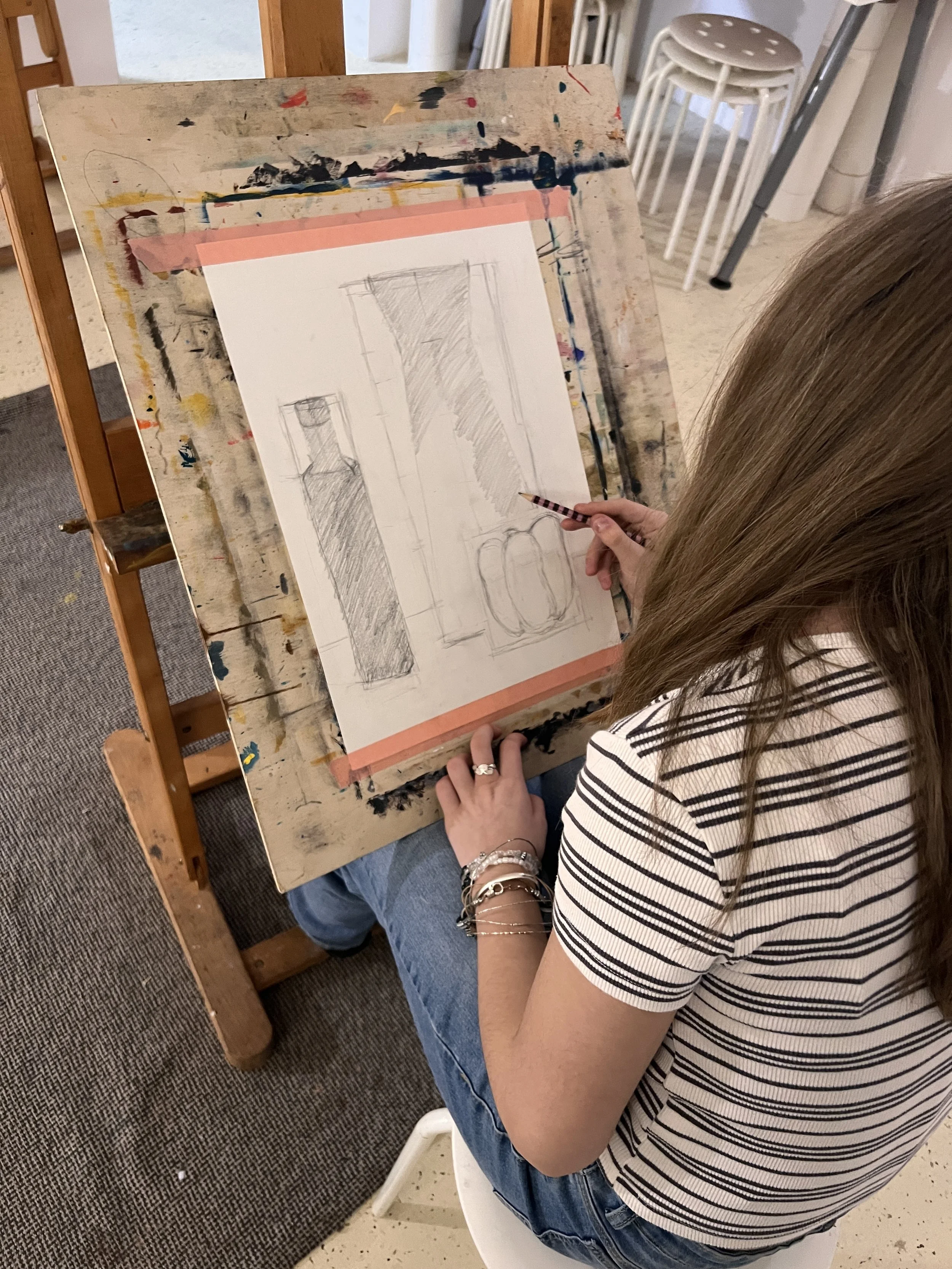 A woman drawing still life sketches of vases and bottles on paper at an art easel in an art studio.