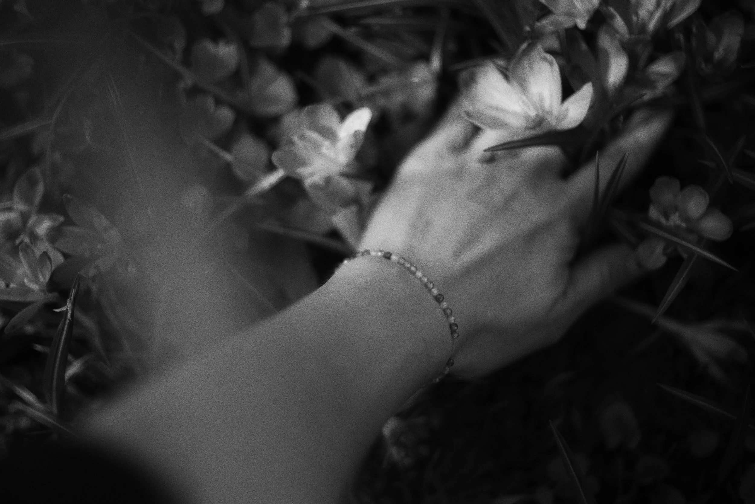 Black and white photo of a person's hand wearing a bracelet, surrounded by flowers and greenery.