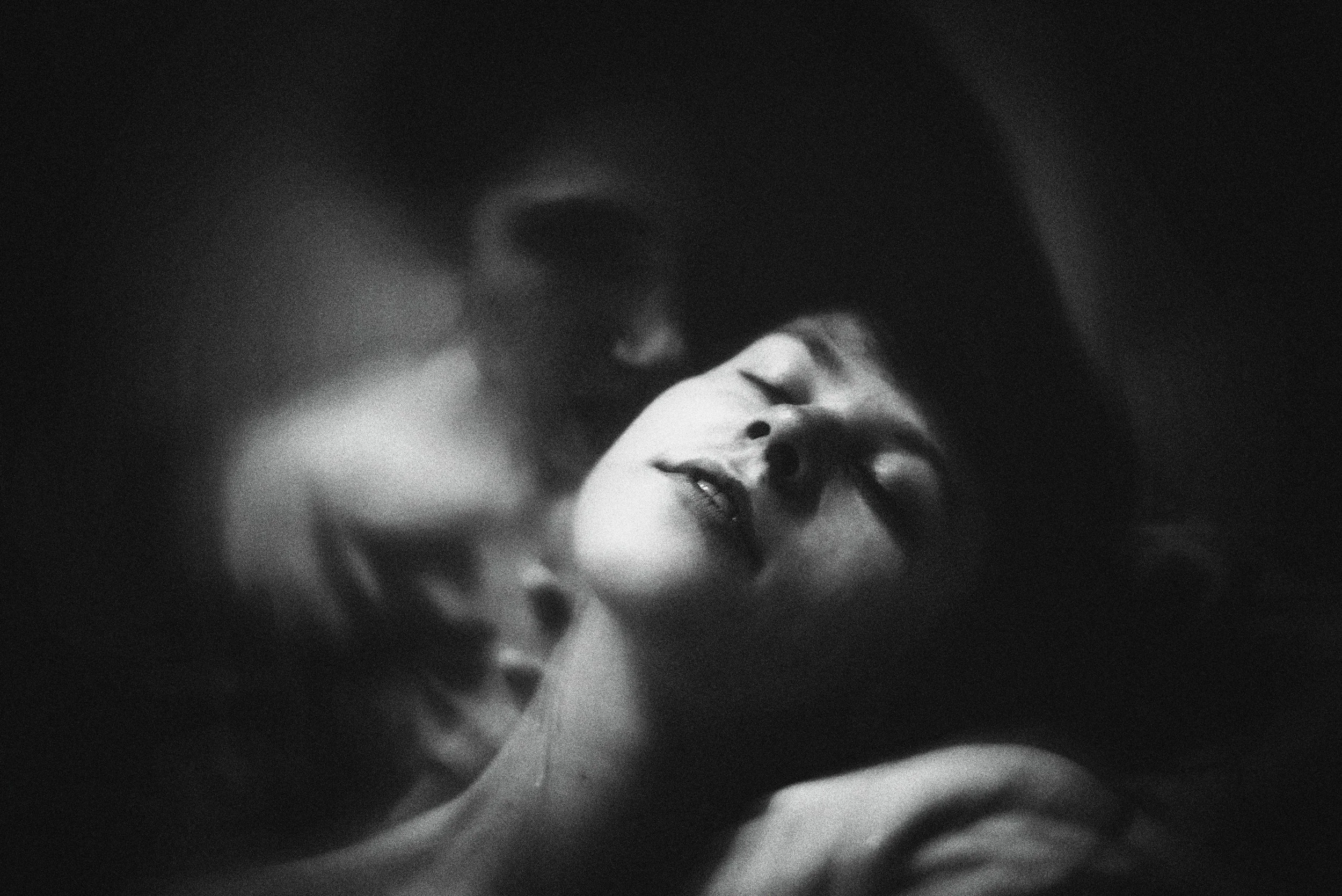 A black and white photo of a woman with closed eyes, with two people close to her face in an intimate pose.