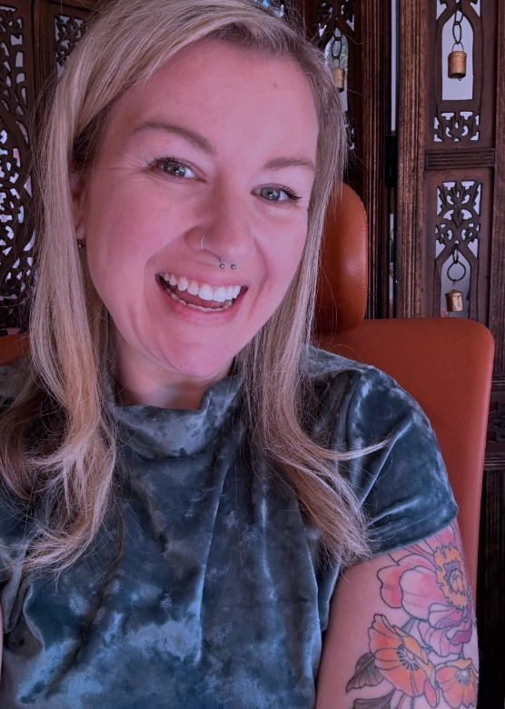 A young woman with blonde hair smiling, showing her teeth, with a septum piercing, seated in front of a wooden decorative screen, wearing a black and gray tie-dye shirt with a floral tattoo on her upper arm.