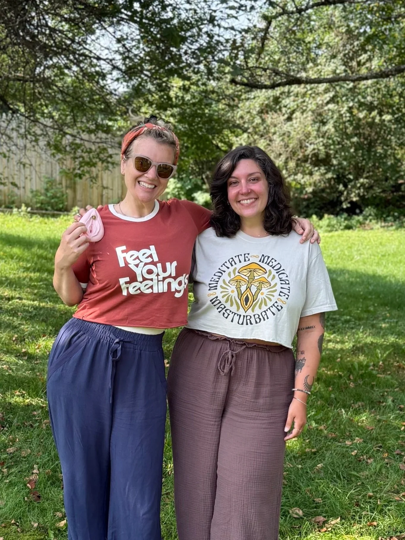 Two women standing on grass with trees in the background, smiling and posing for a photo. One woman wears sunglasses, a red t-shirt, and blue pants, holding a pink item. The other woman has dark curly hair, a light-colored t-shirt with a circular design and text, and brown pants.