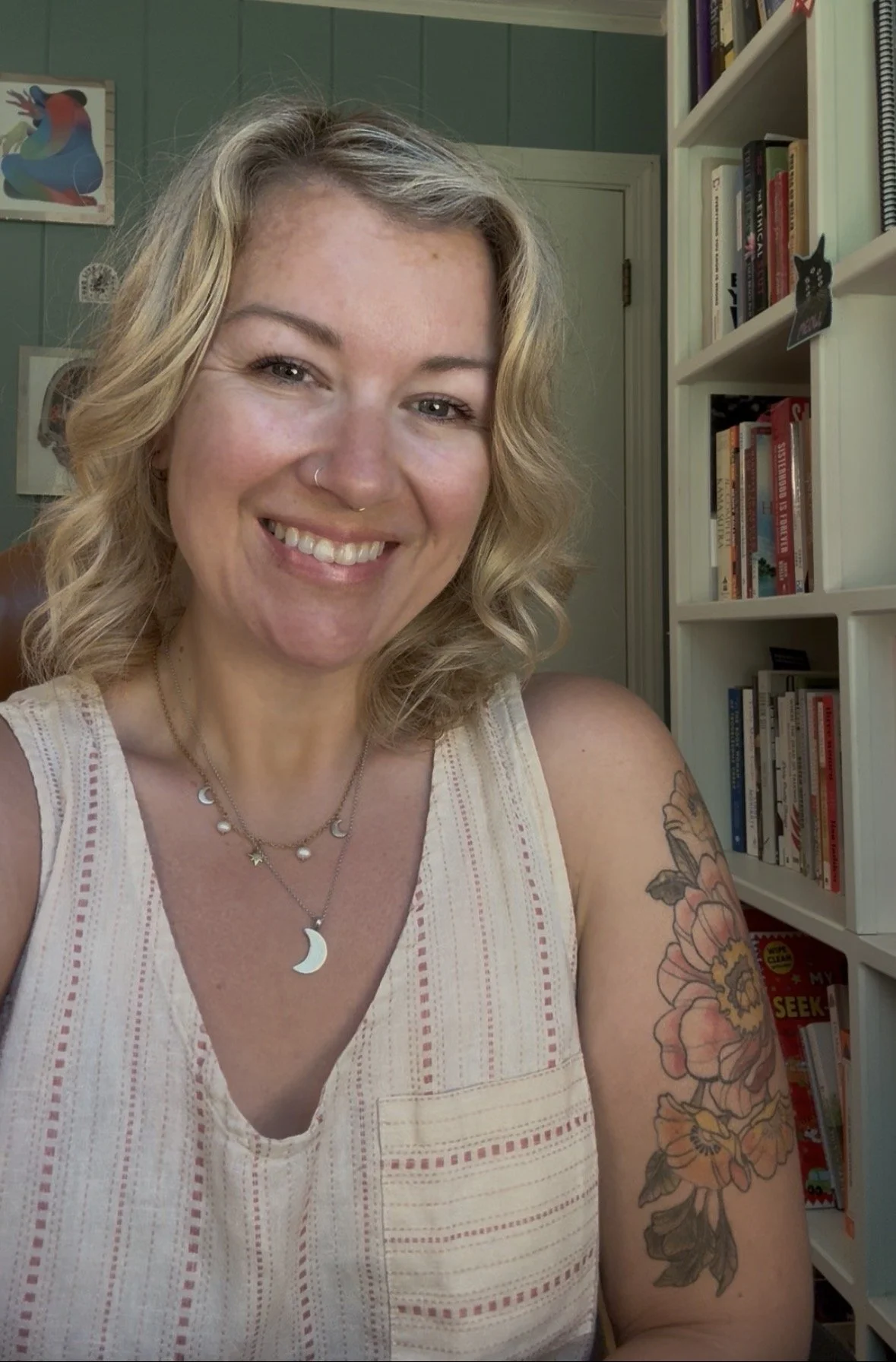 A woman with blonde hair smiling, wearing layered necklaces with moon and star charms, and a sleeveless beige top with red stitching. She has floral tattoos on her left arm. Behind her are bookshelves filled with books, and a wall with artwork, including a colorful bird painting.