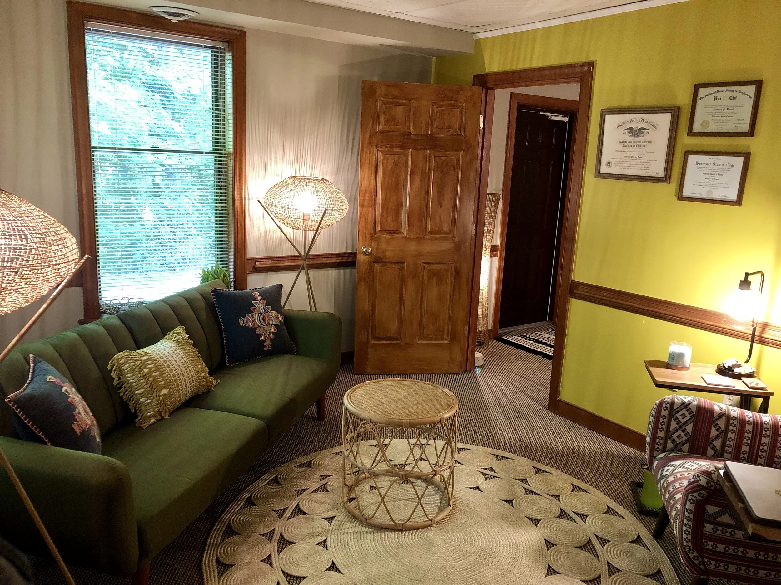 A cozy living room with a green velvet sofa adorned with colorful patterned pillows, a rattan coffee table, and a woven lamp standing next to a window with blinds. The right wall is painted yellow and decorated with framed diplomas. There is a doorway leading to another room, and a side table with various items on it.
