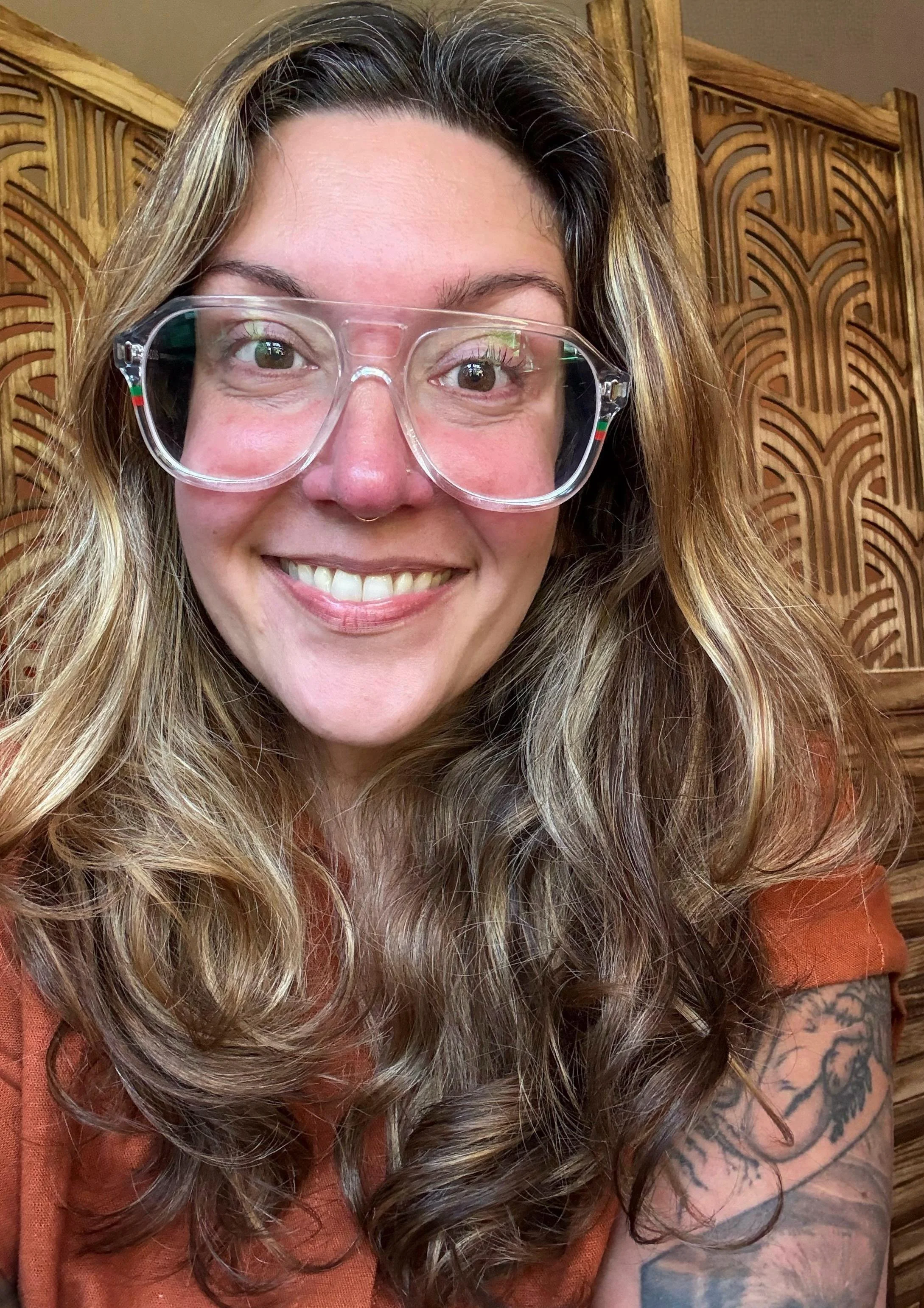 A close-up photo of a woman with wavy blonde hair, wearing clear glasses with a small flag detail on the temples, smiling, with a wooden decorative background and a tattooed arm visible.