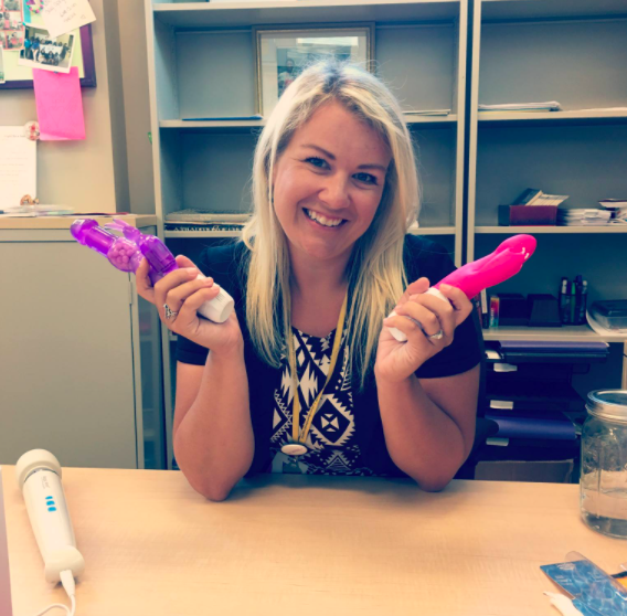 Woman smiling at desk holding two vibrators, one purple and one pink.