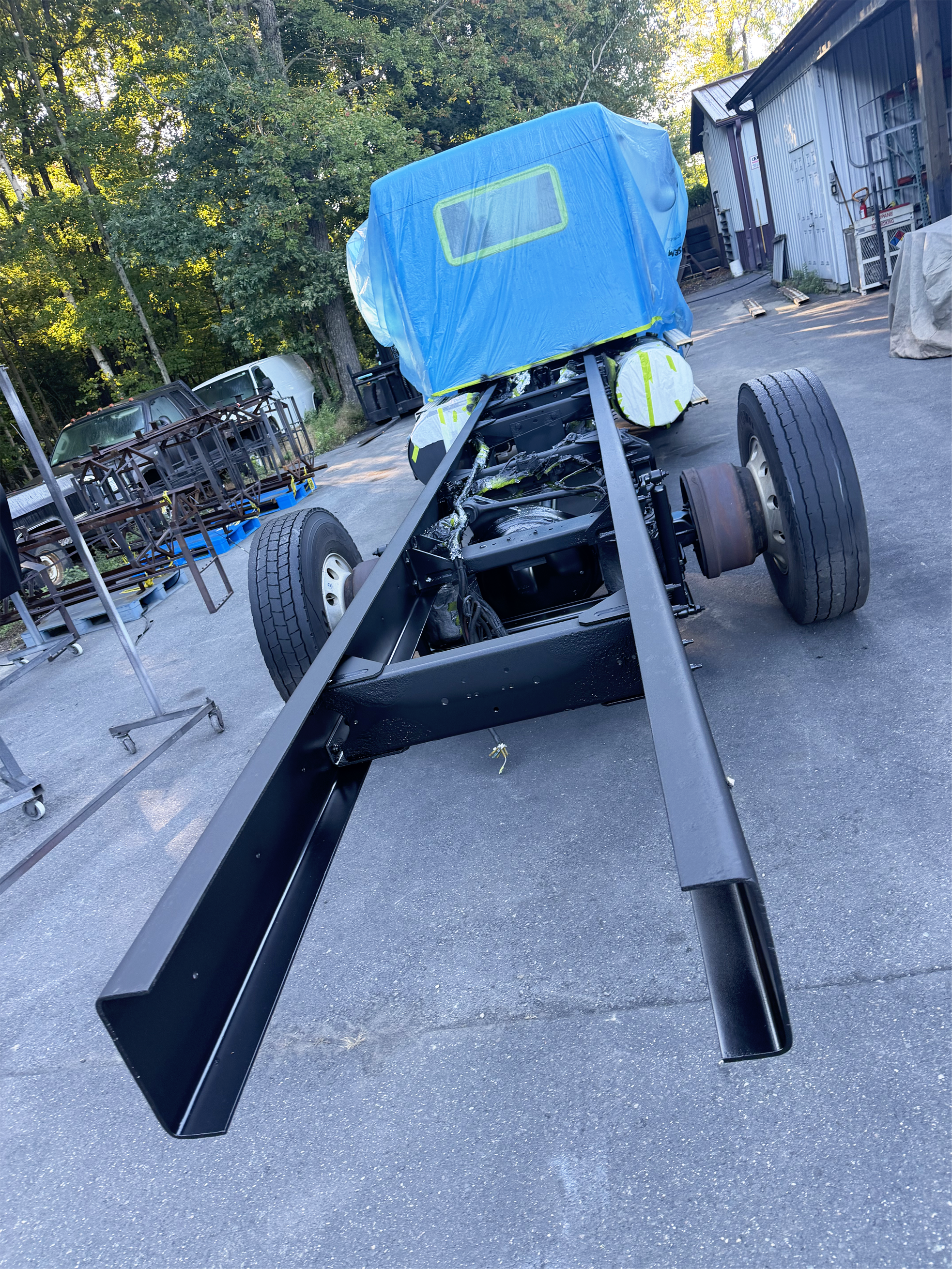 Partially assembled vehicle chassis with four wheels and a blue protective cover outside a workshop