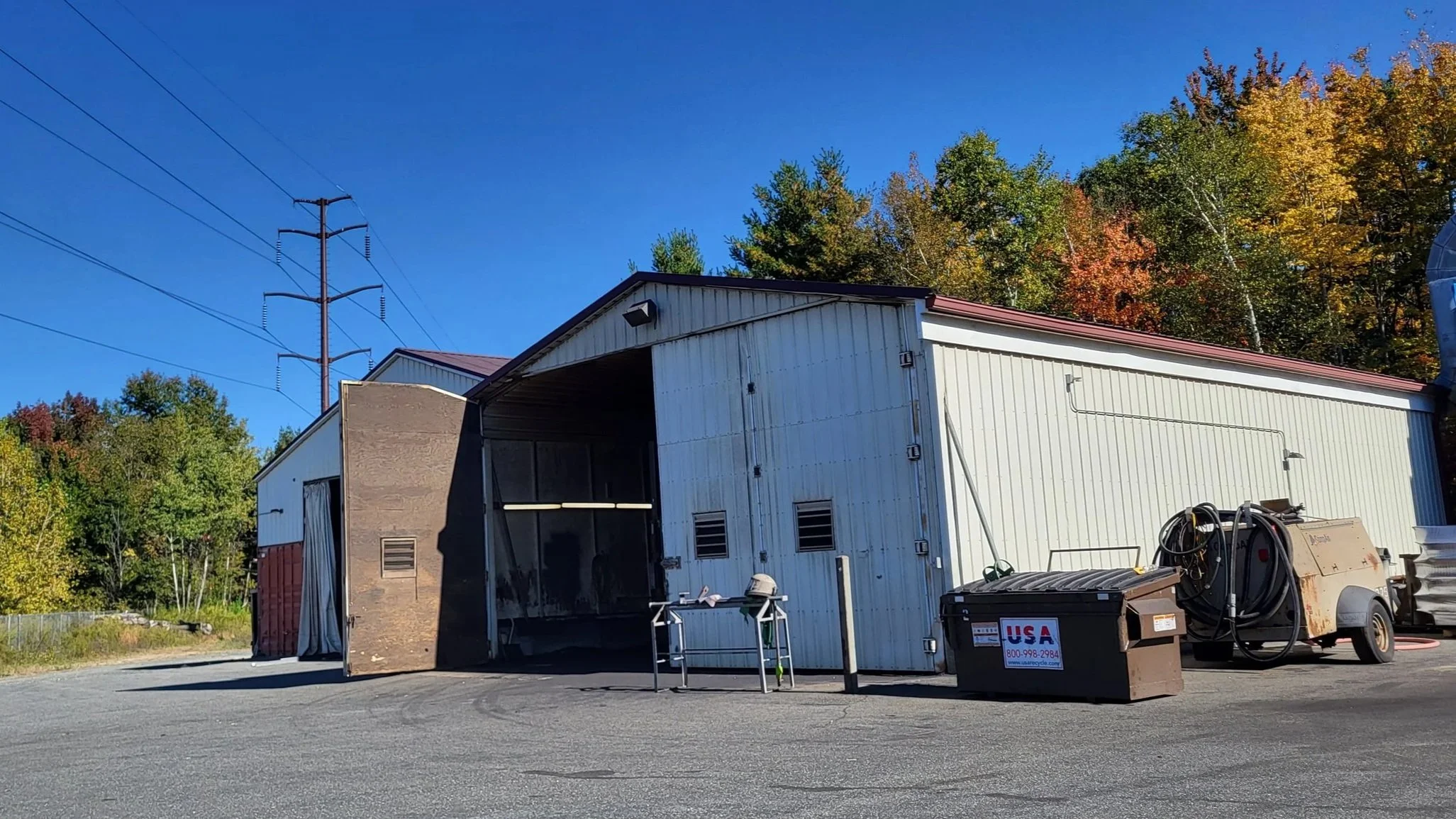 Industrial maintenance building with open garage, utility poles, dumpsters, and equipment trailer against autumn trees