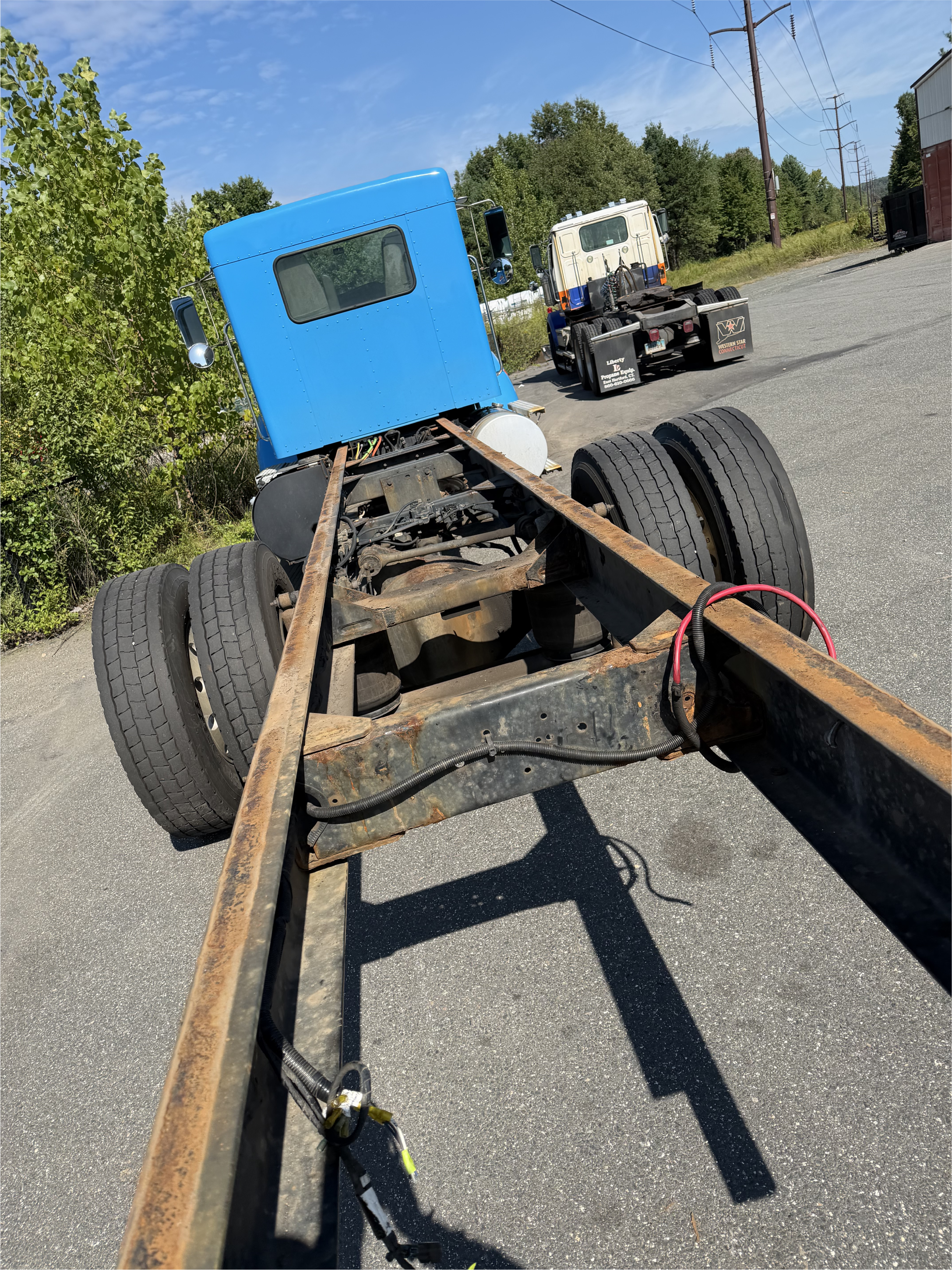 Blue semi-truck chassis with exposed rusty frame parked beside a cream-and-blue truck in an industrial lot