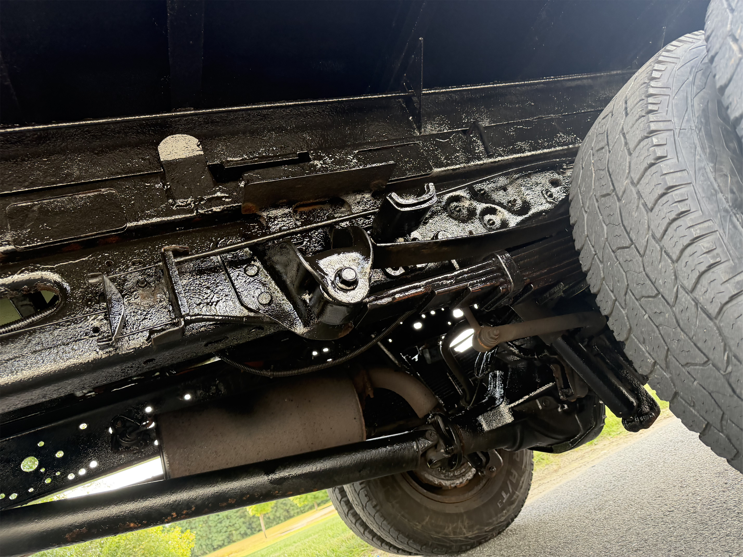 Underside of a black off-road vehicle showing the frame, suspension, axle, exhaust, hitch components, and rugged tires.