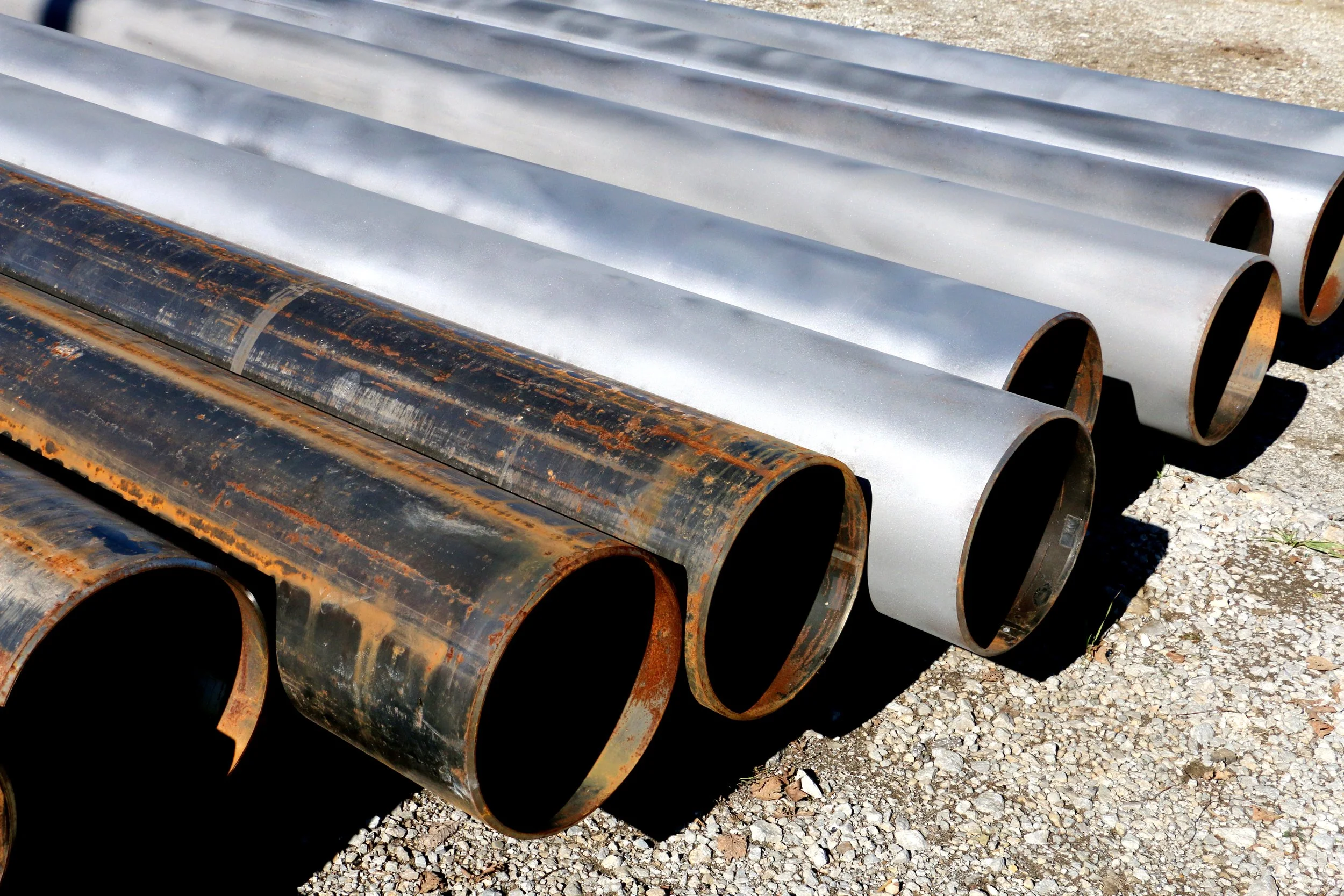 Large rusted and galvanized steel pipes stacked outdoors on gravel