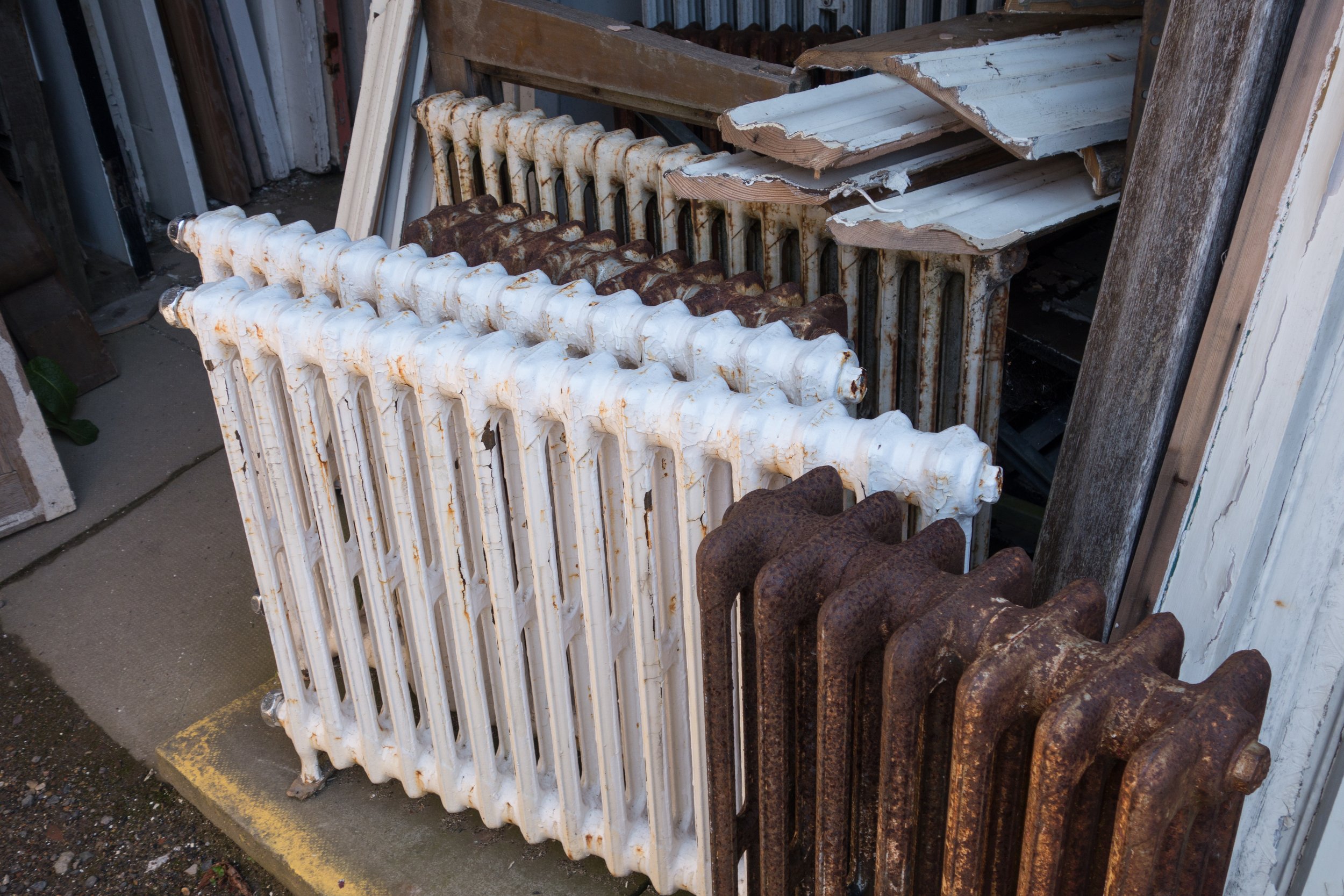 Stacked vintage cast-iron radiators, some painted white and others heavily rusted, beside salvaged wooden trim.