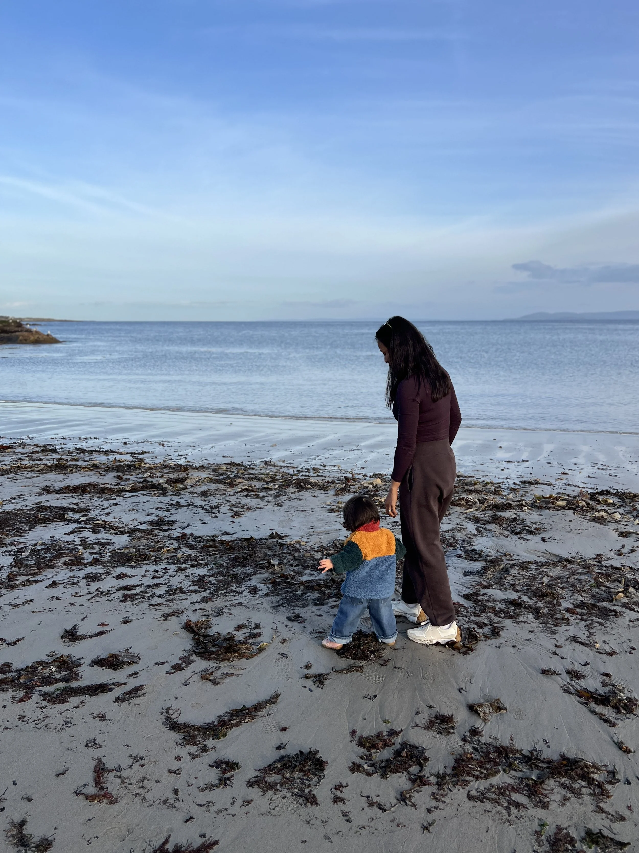 A woman and a young child walk along the beach near the water, with sandy shore and scattered seaweed in the foreground, and clear blue sky with a few clouds in the background.