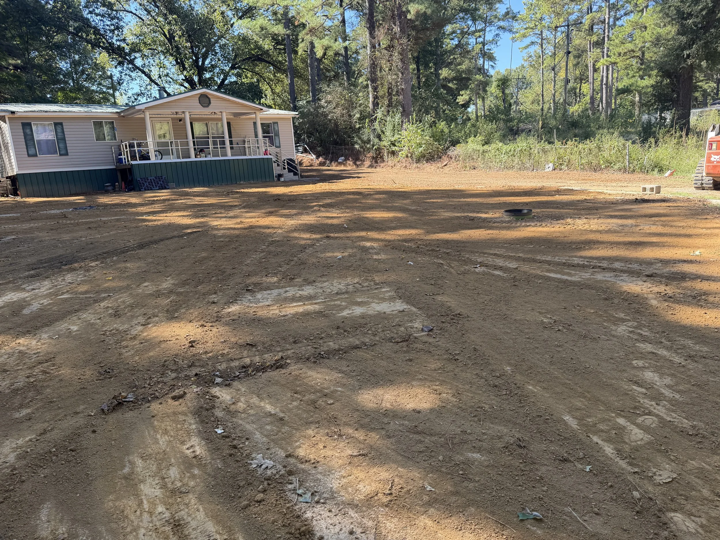 A cleared patch of land with mostly dirt, some scattered leaves, and tire marks, bordered by a house with a porch and lush green trees in the background.