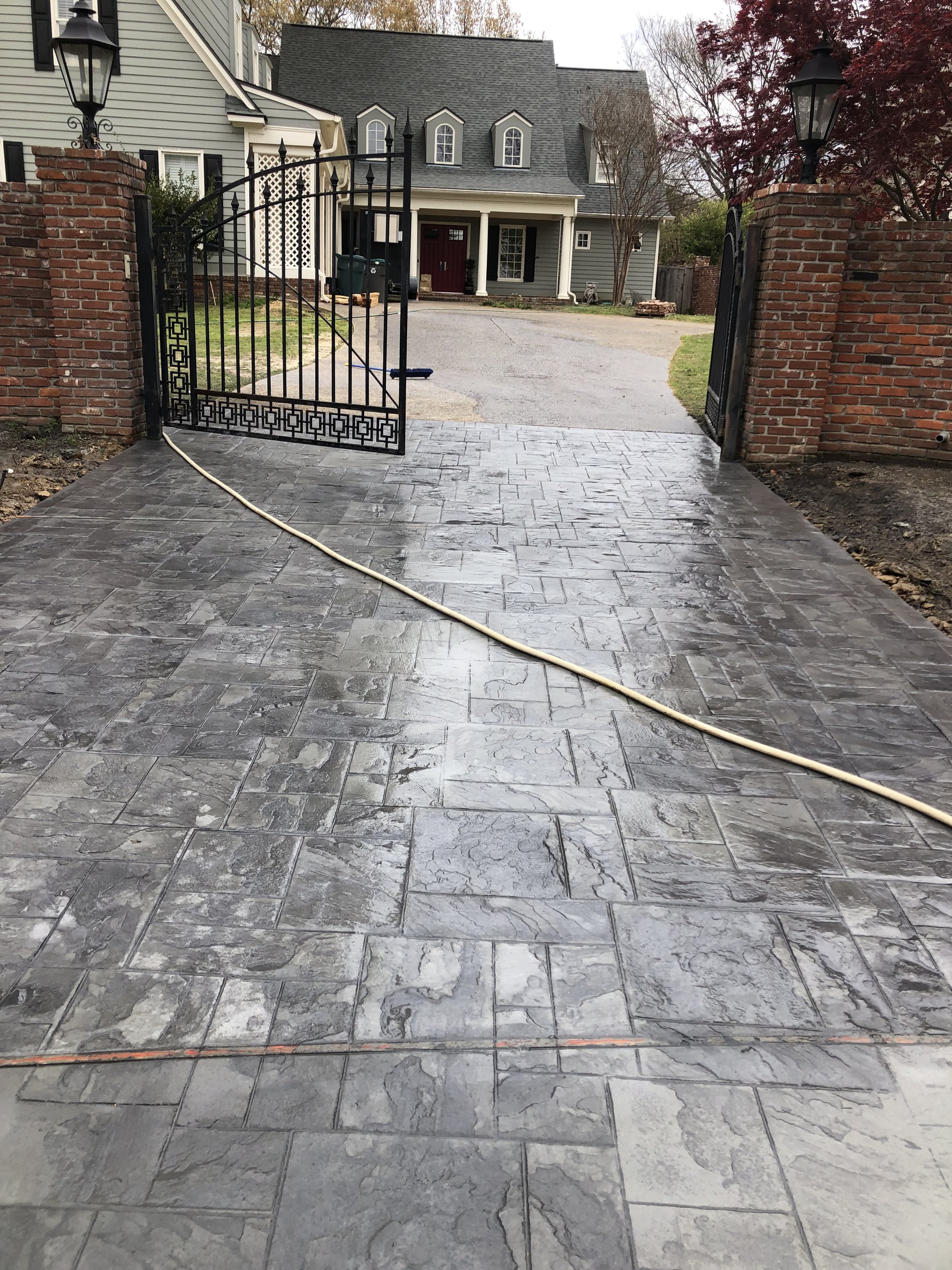 Freshly installed gray stone patio in front of a house with a black wrought iron gate and brick pillars, a garden hose lying on the surface.