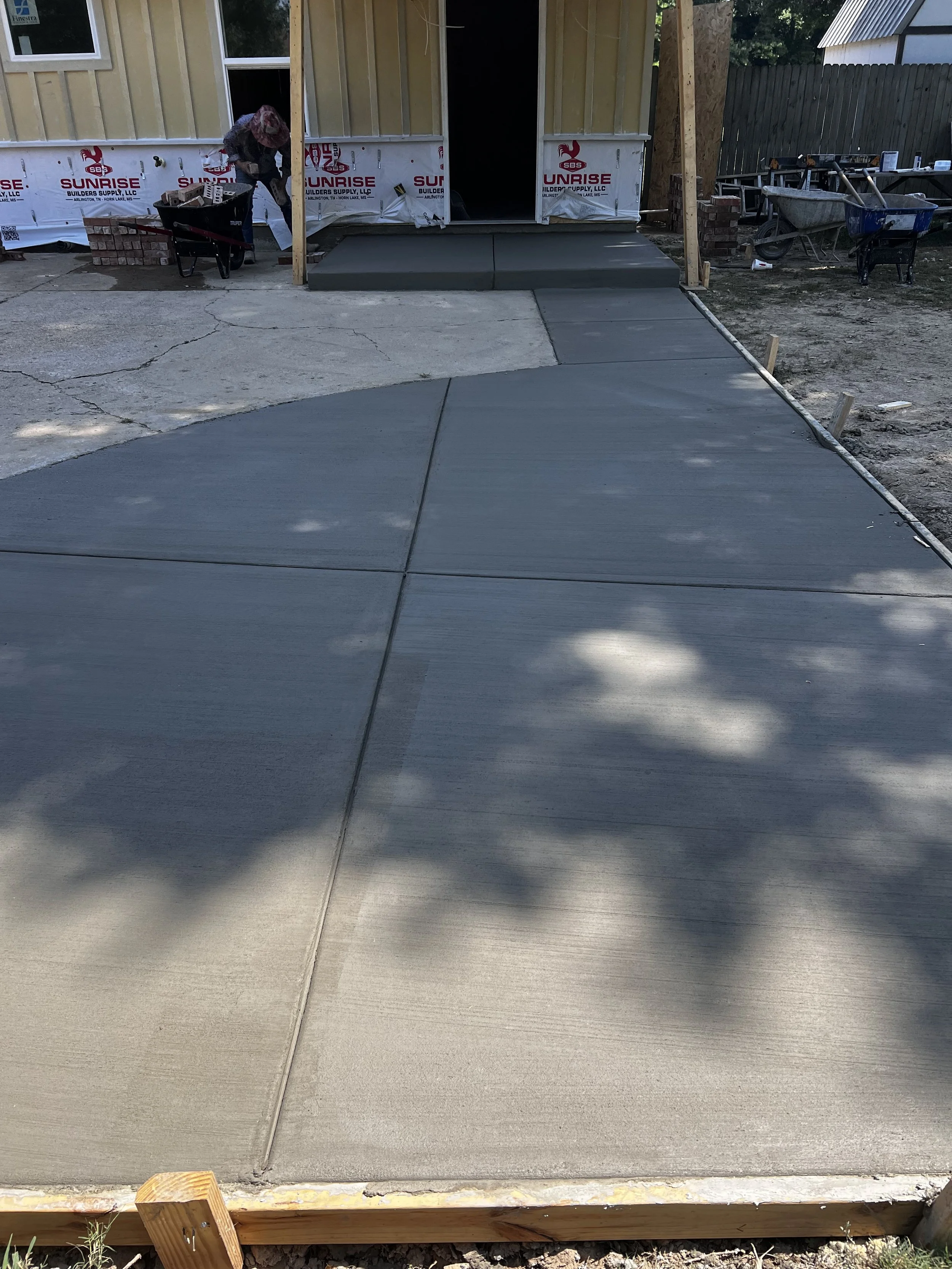 Newly poured concrete sidewalk leading to the front door of a house under construction, with construction tools and materials around.