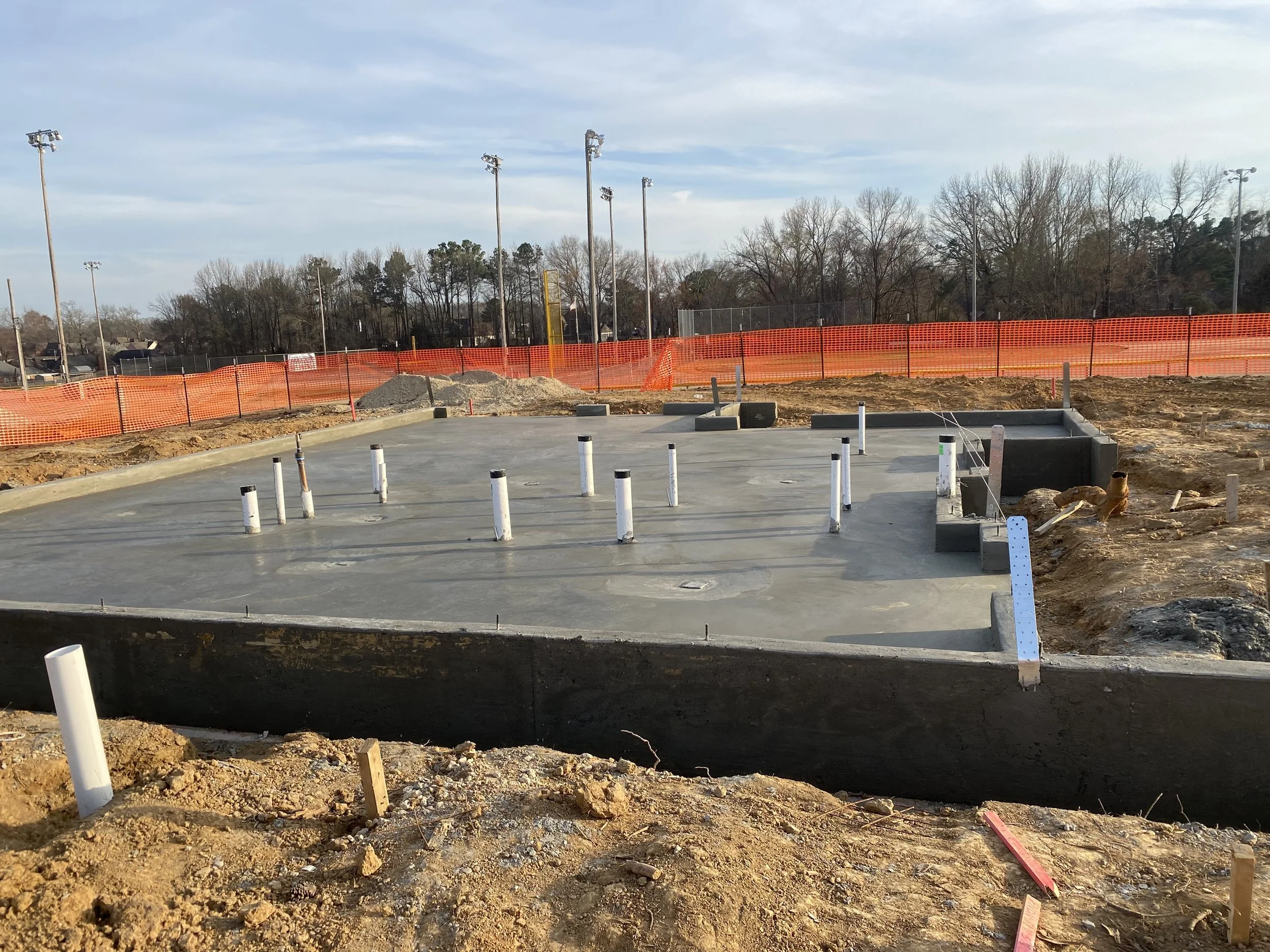 Construction site with a freshly poured concrete slab, orange safety fencing, and several utility pipes sticking out of the ground. There are tall outdoor lights, leafless trees in the background, and a partly cloudy sky.