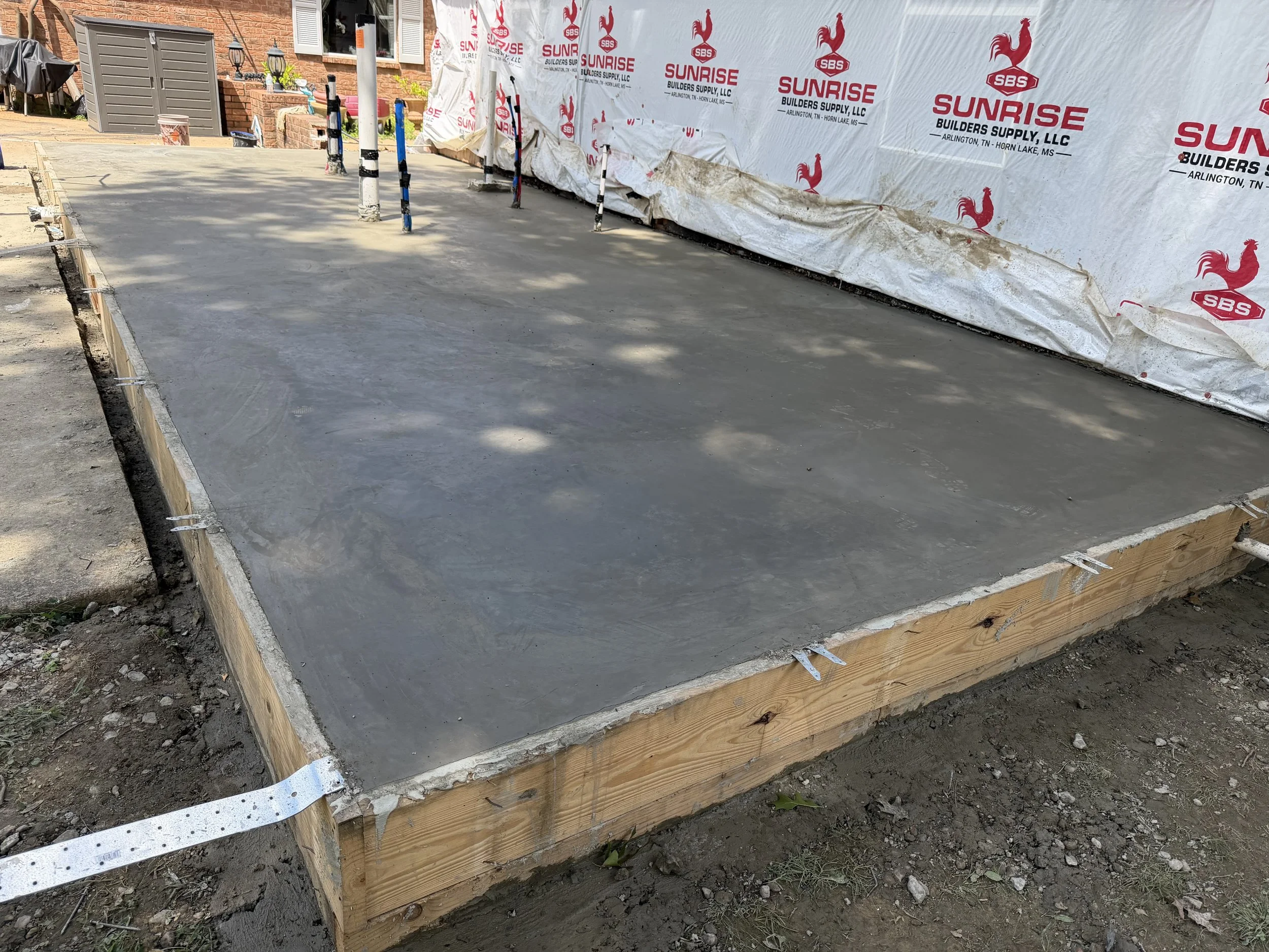 Concrete slab poured on a wooden framed foundation at a residential construction site, with construction dirt and some grass in the foreground, and a house with brick exterior and a white window in the background, along with building materials and a covered outdoor grill.