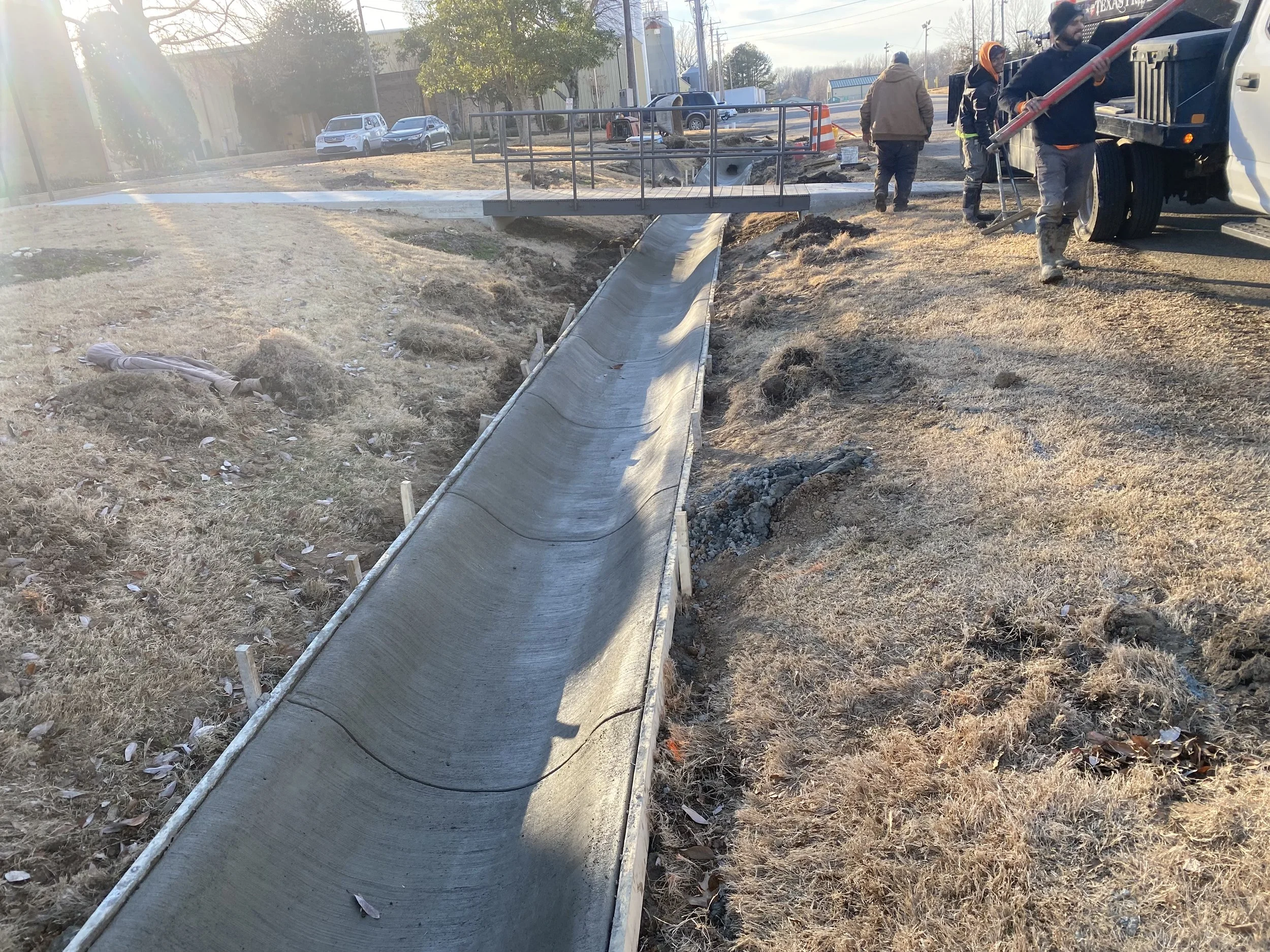 Construction workers installing a concrete sidewalk with temporary wooden framing, nearby a small bridge, on a grassy area.