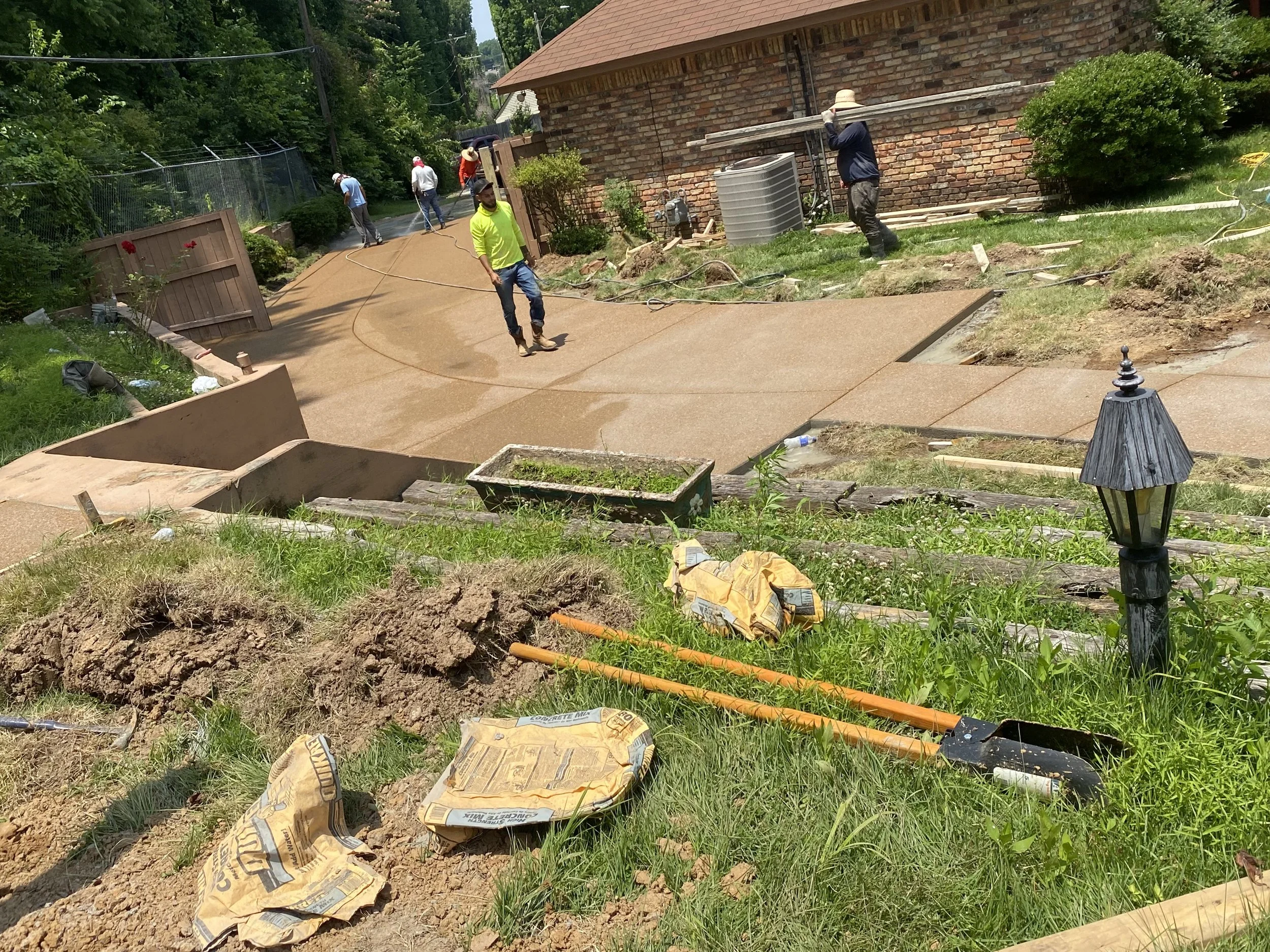 Construction workers pouring concrete on a driveway in front of a brick house. The scene includes tools, bags of concrete mix, and freshly poured concrete.