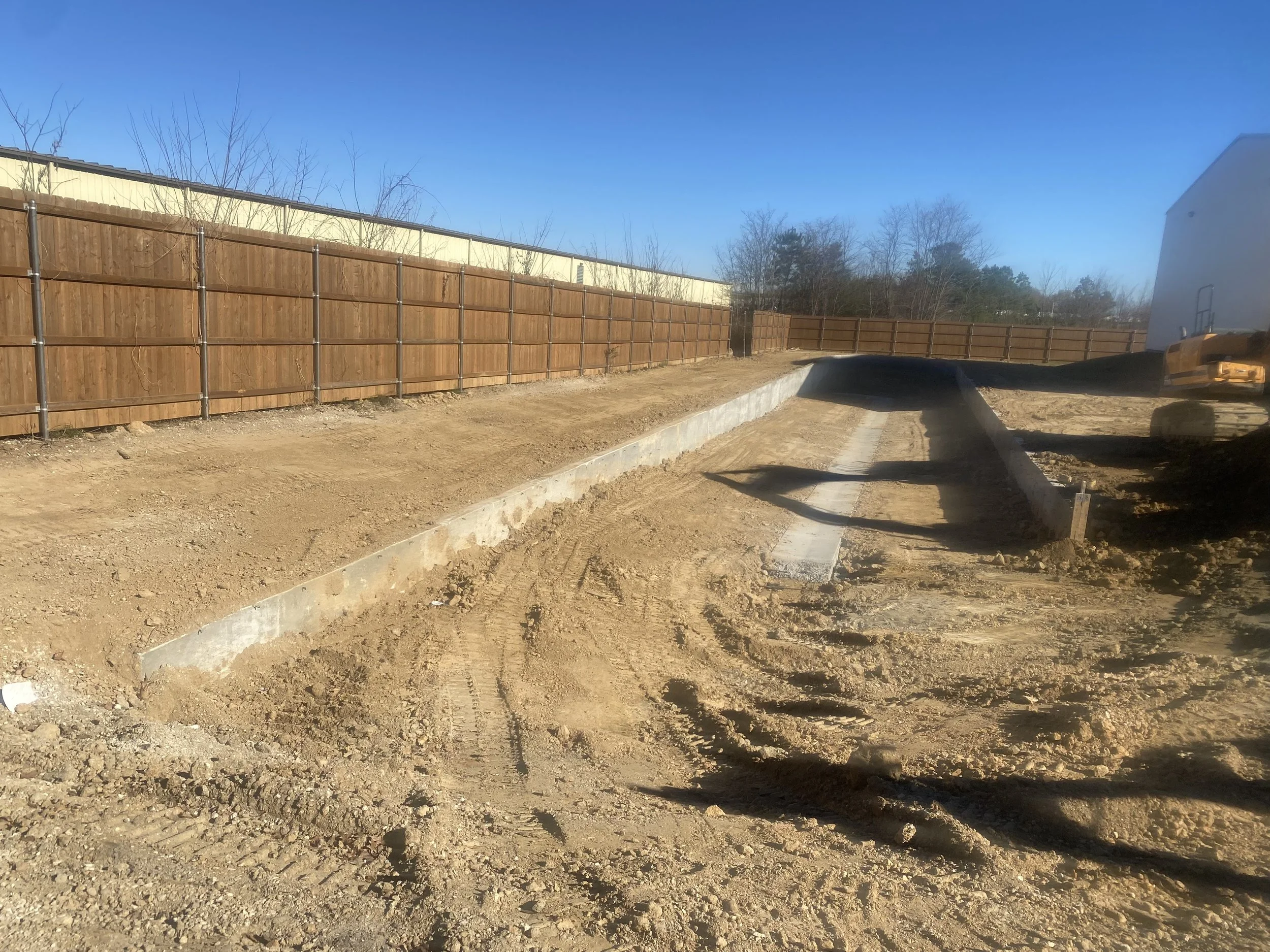 Construction site with a dirt ground, wooden fence, and a yellow excavator in the background under a clear blue sky.
