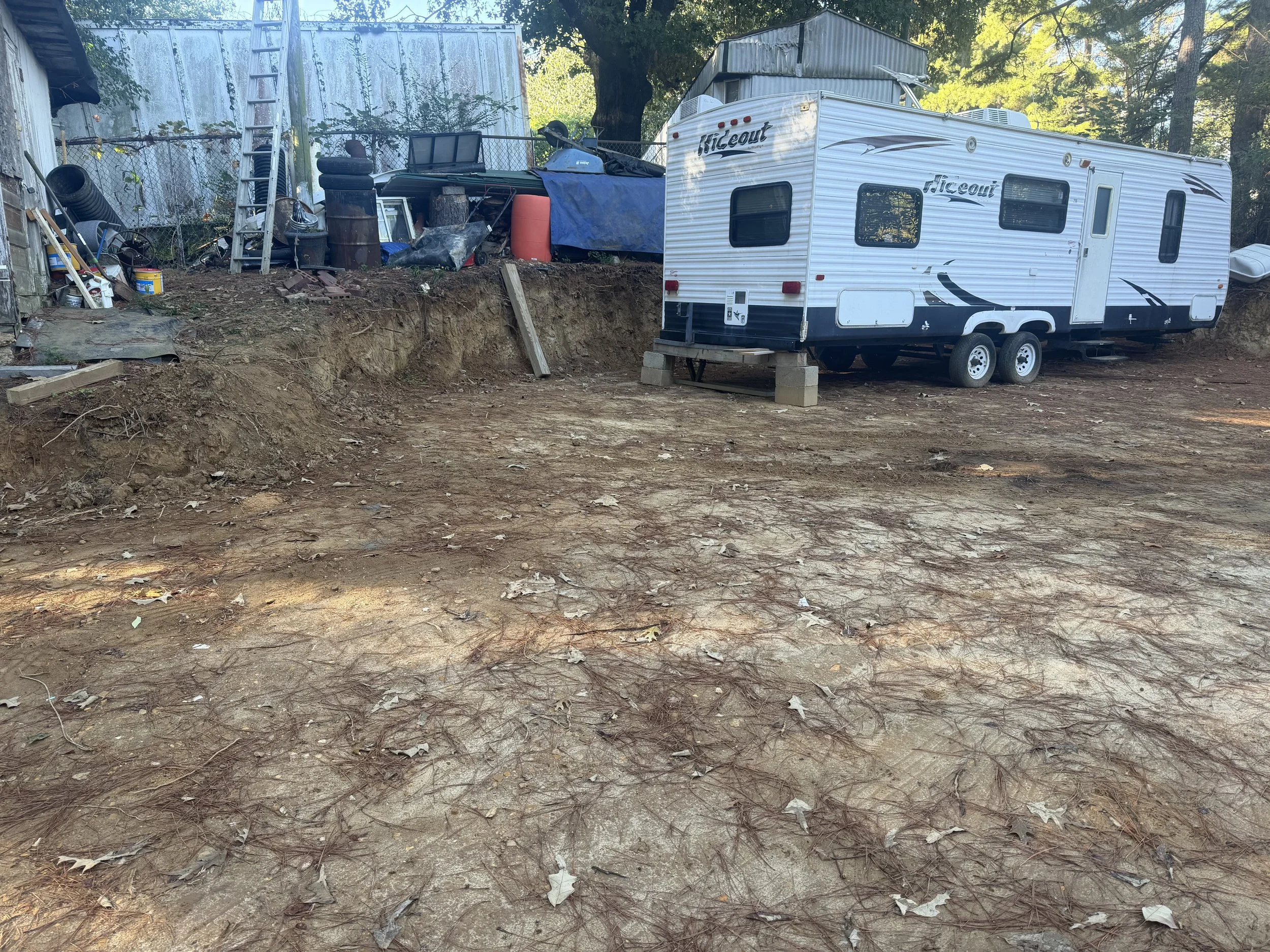 An RV trailer parked on a dirt lot with scattered leaves, next to a fenced area with various items, tools, and a ladder, under trees with green leaves.