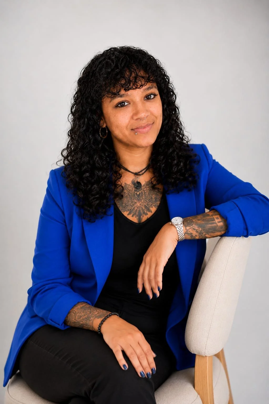 Junge Frau mit Tattoos, Armbanduhr, blaurem Blazer und schwarzen Kleidung sitzt auf einem Stuhl vor hellem Hintergrund.