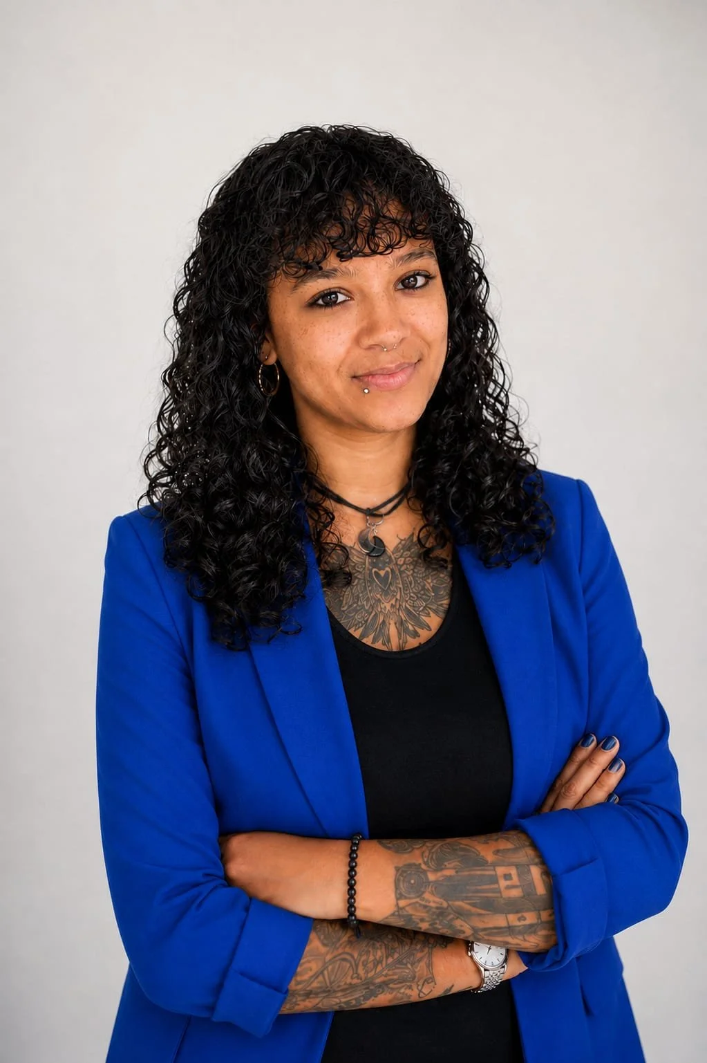 Eine Frau mit dunklen, Lockigen Haaren, Tattoo auf Brust und Armen, in einer blauen Jacke und schwarzen Top, steht vor einem weißen Hintergrund.