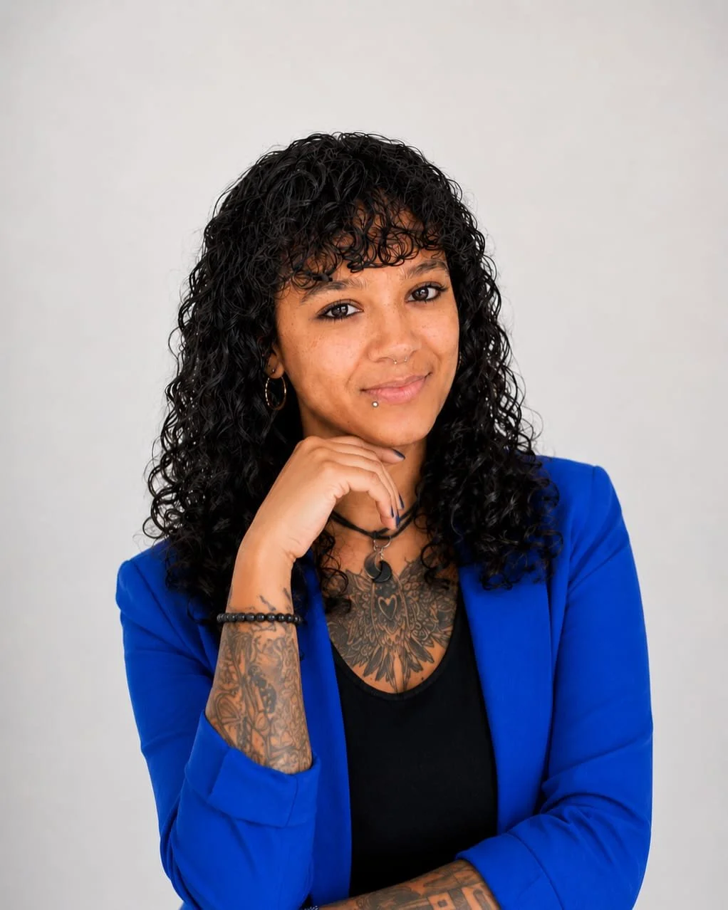 Eine Frau mit schwarzen, lockigen Haaren, Tattoos an Armen und Brust, trägt eine blaue Jacke und hat Piercings im Gesicht, posiert vor einem neutralen Hintergrund.