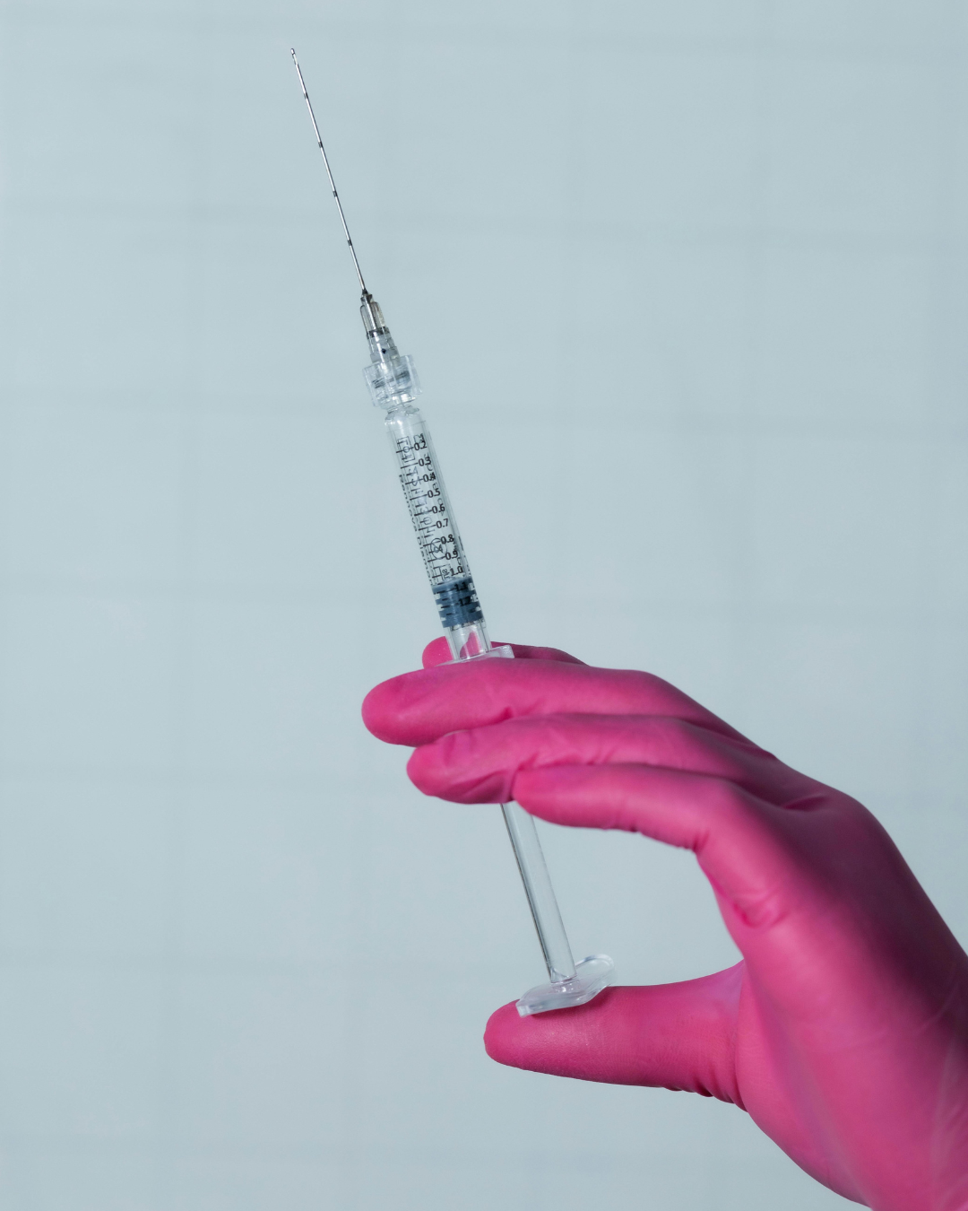 A hand wearing a pink glove holding a syringe against a light background.