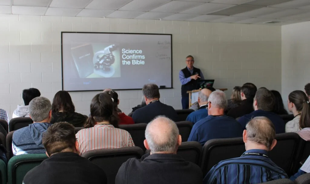 A man giving a presentation to a seated audience in a room with a white brick wall. The screen behind him displays an image of a microscope and a Bible with text reading 'Science Confirms the Bible'.