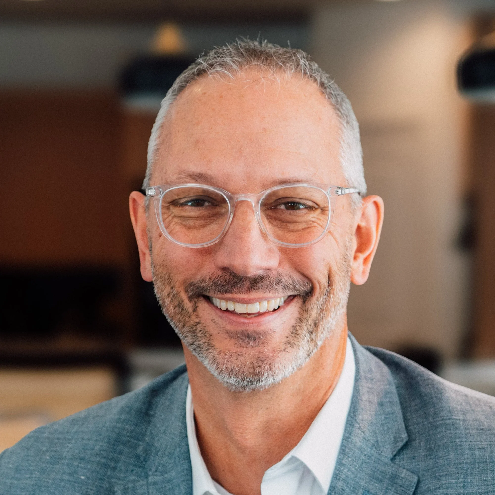 Close-up of a smiling man wearing clear glasses and a business suit with a light blue shirt. He has short gray hair and a trimmed gray beard. Featuring Mark Vroegop