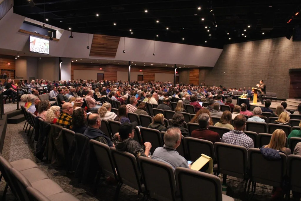 Large audience seated in a conference hall, attending a speaker's presentation on stage.