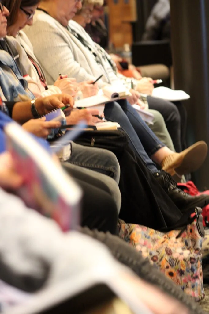 Group of people sitting in a row, taking notes or reading, in a conference or seminar setting.