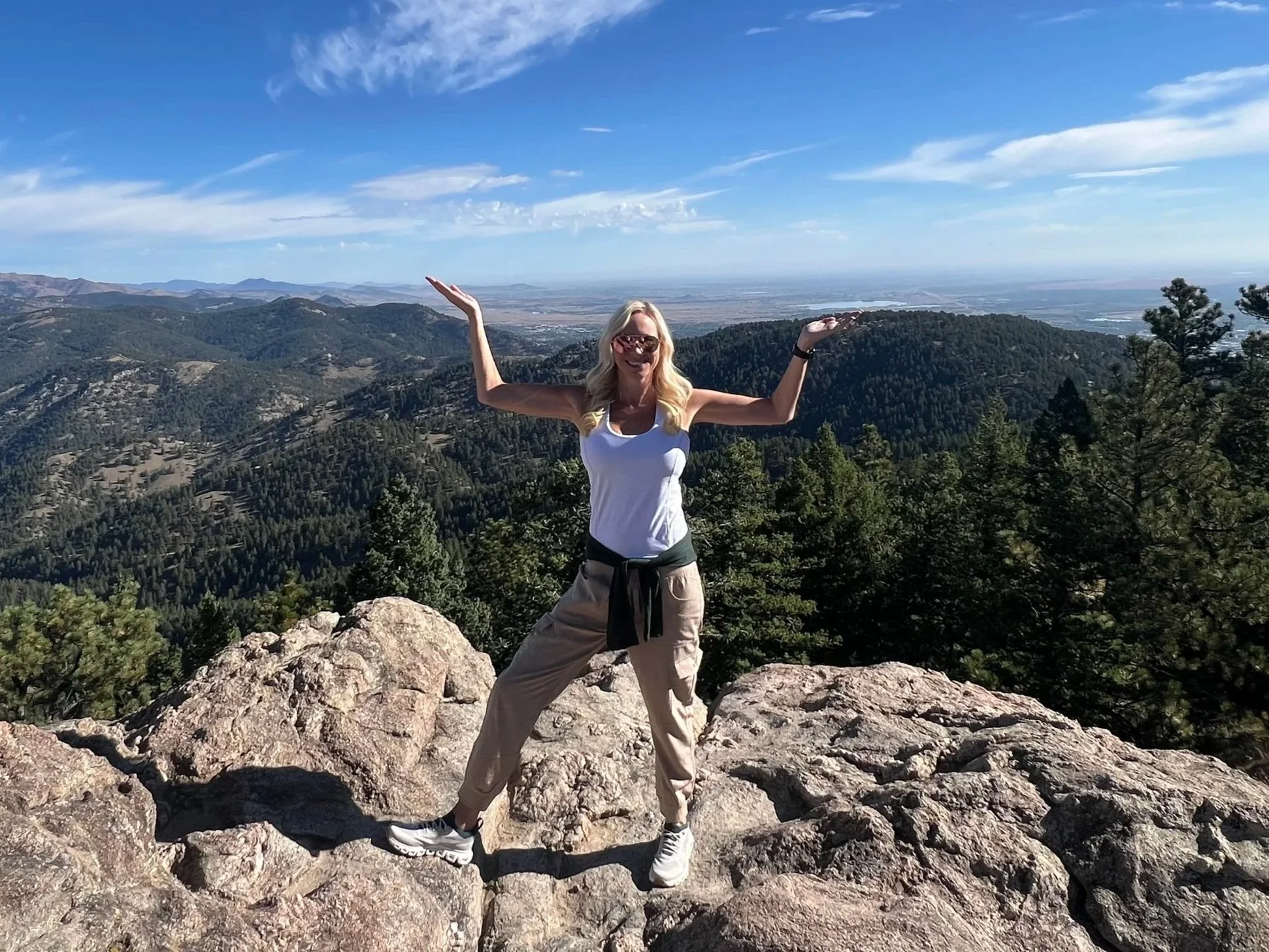 A woman with blonde hair and sunglasses standing on a rocky mountain with her arms raised in front of a scenic landscape of valleys, hills, and forests under a blue sky with clouds.