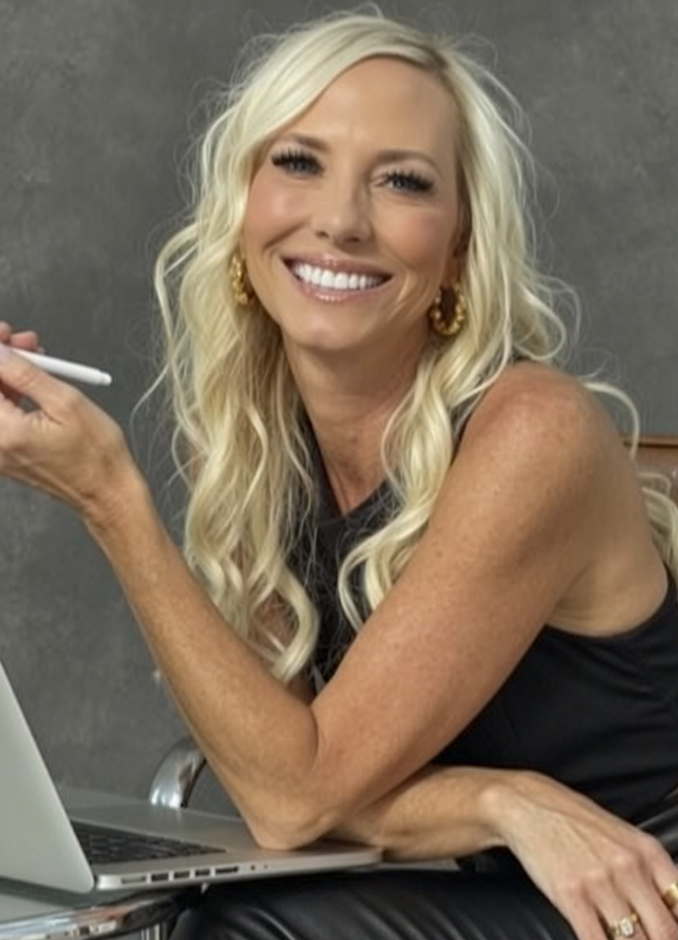 A smiling woman with long blonde hair, wearing a black sleeveless top, gold earrings, and holding a white pen, sitting at a desk with a laptop in front of her.