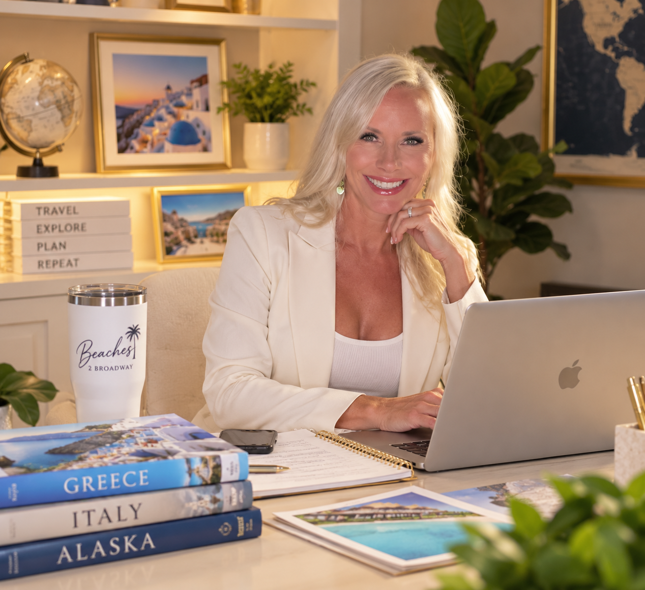 A woman with blonde hair smiling sitting at a desk with travel books, a laptop, and a tumbler that says 'Beaches 2 Broad Way.' The background features framed photos of Greece, a globe, and a plant.