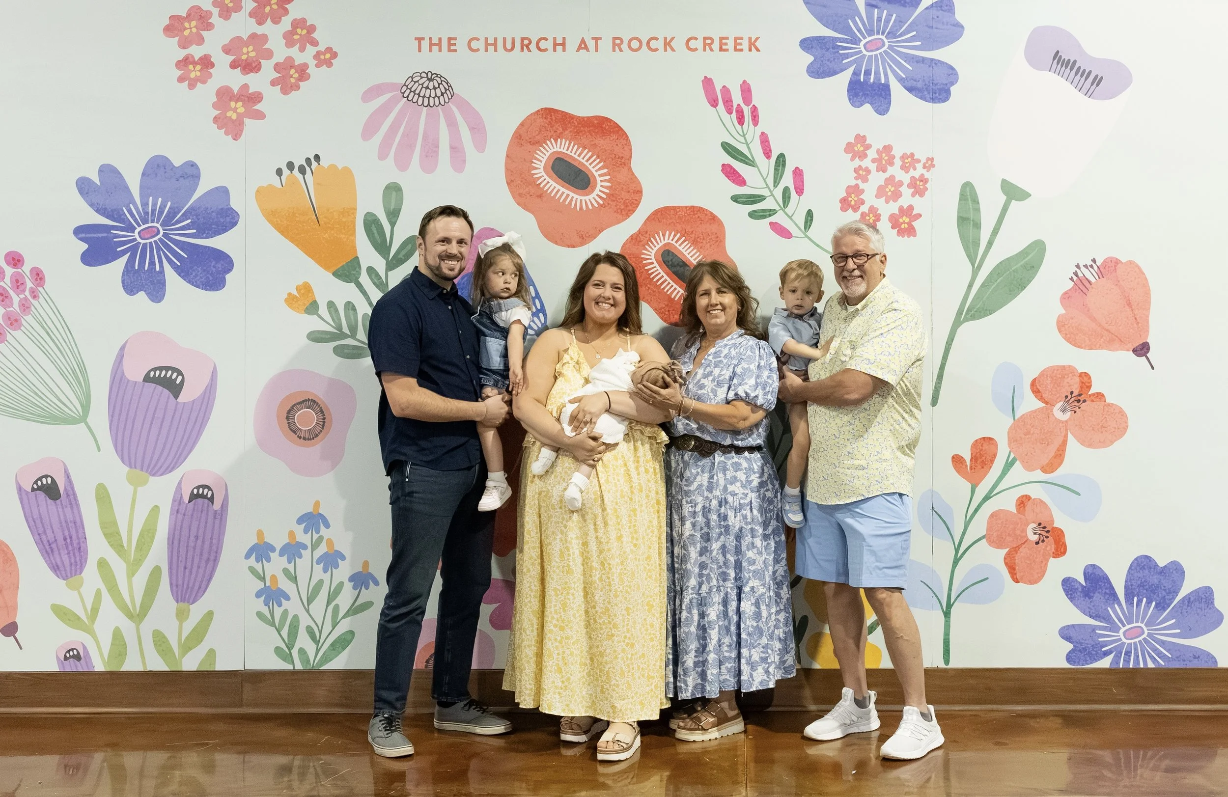 Family group standing in front of a colorful floral mural with the text 'The Church at Rock Creek.' The group includes three adults, two children, and a baby, with all smiling and posing for the photo.
