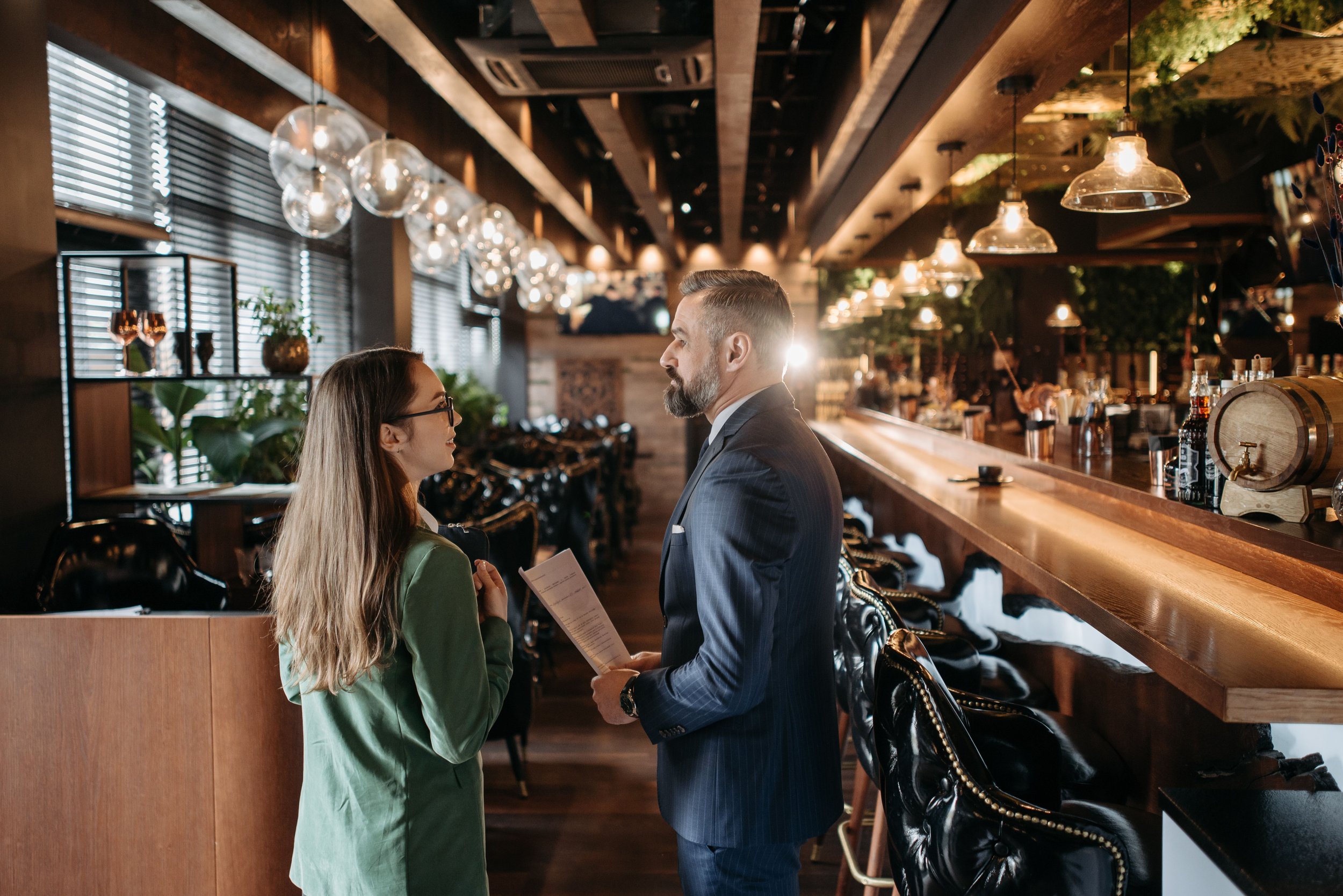 A man in a suit and a woman in a green blazer are conversing at a bar inside a warmly lit restaurant or bar, with decorative glass lamps hanging from the ceiling.