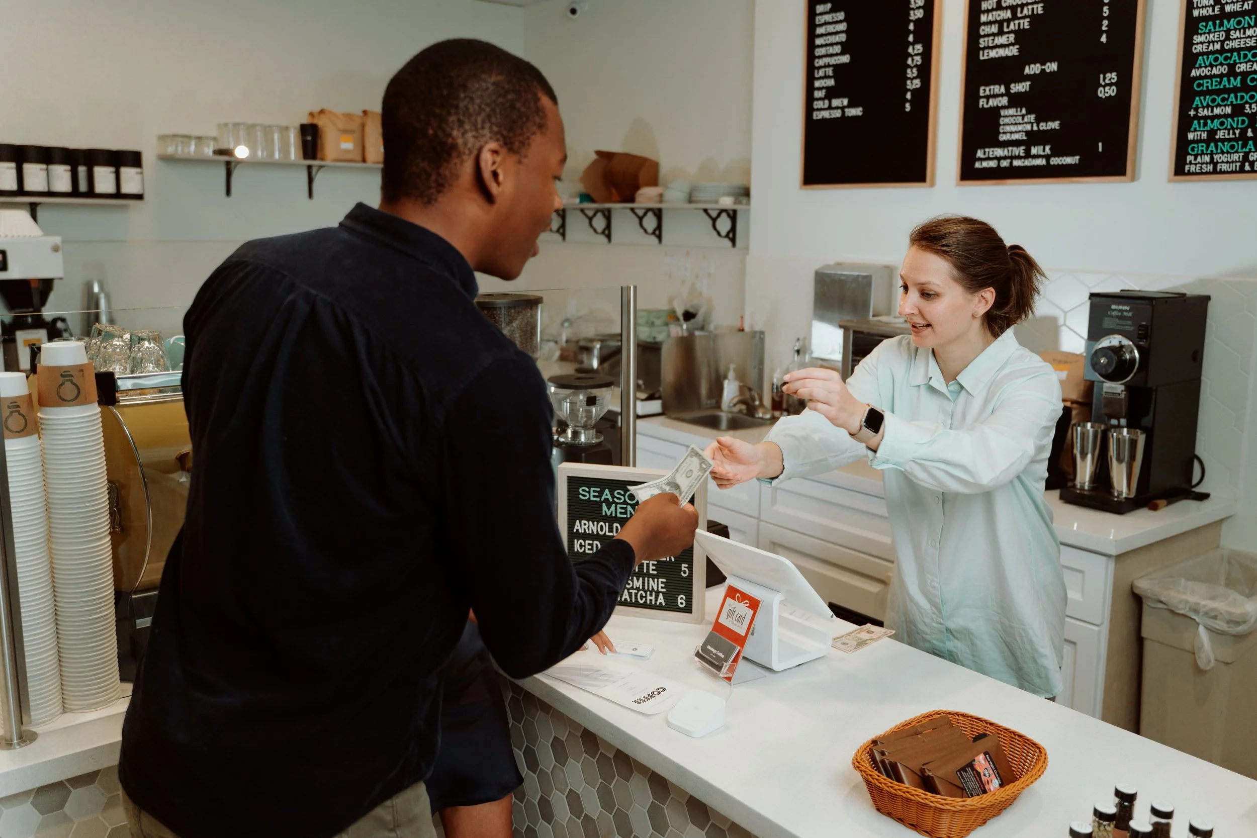 A man paying with cash at the counter of a coffee shop, while a female employee hands him the change. The counter has a display of coffee options and a basket of gift cards.