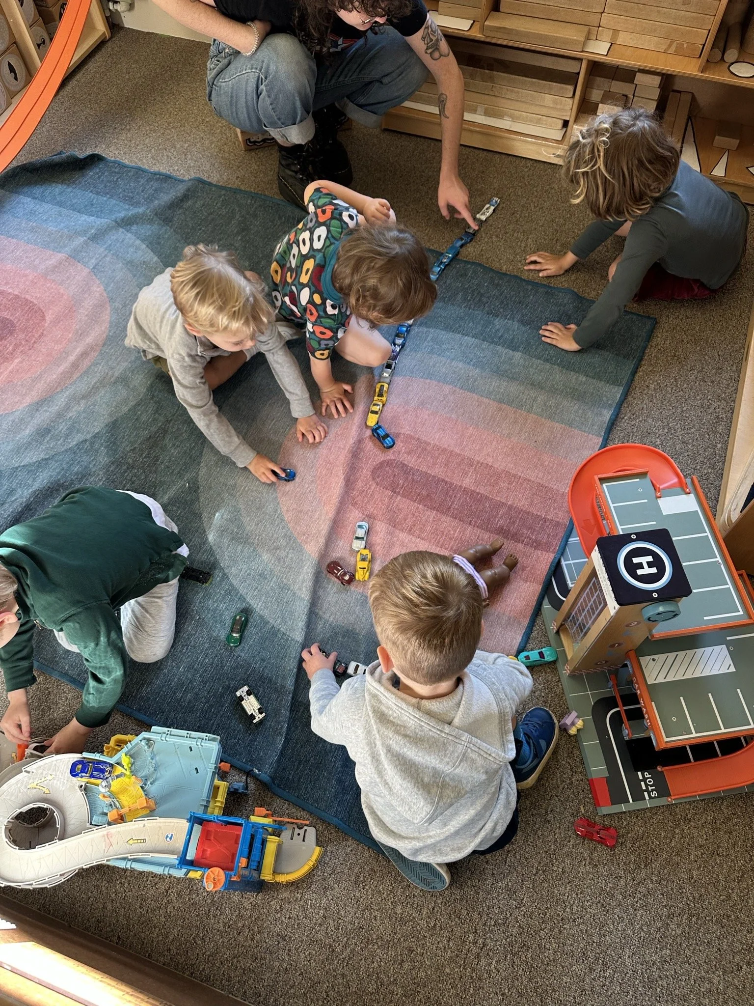Children playing on a colorful rug at Youngset Preschool with toy cars and a play hospital set nearby, supervised by a volunteer caregiver crouching down.