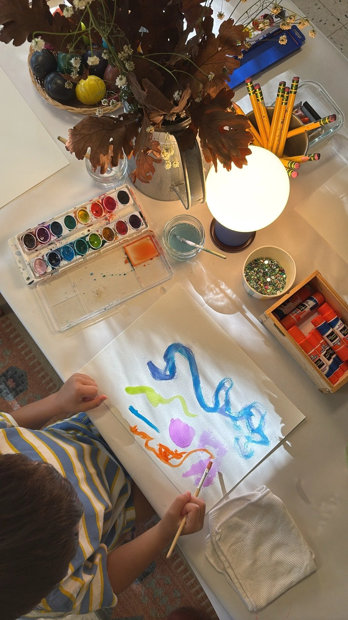 Children painting with watercolors and drawing tools on paper at a table decorated with fall leaves, a basket of small pumpkins, and art supplies including glue sticks, paint, and glitter.