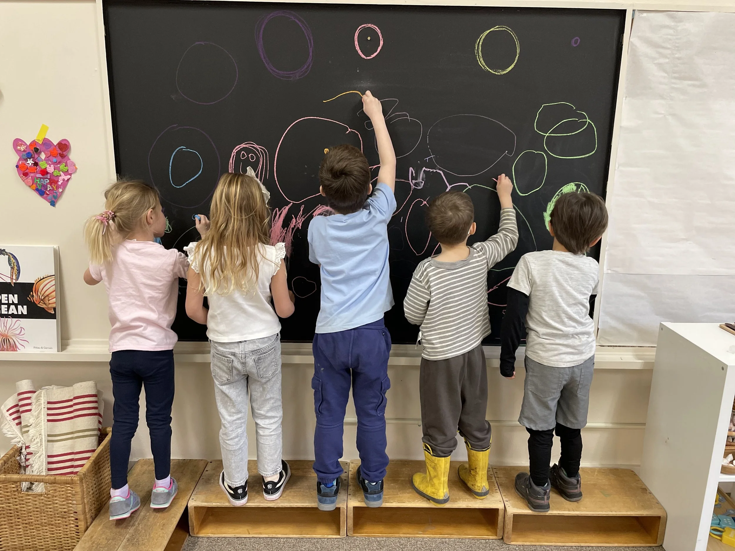 Six children drawing on a blackboard with colorful chalk, standing on a wooden platform in a classroom.