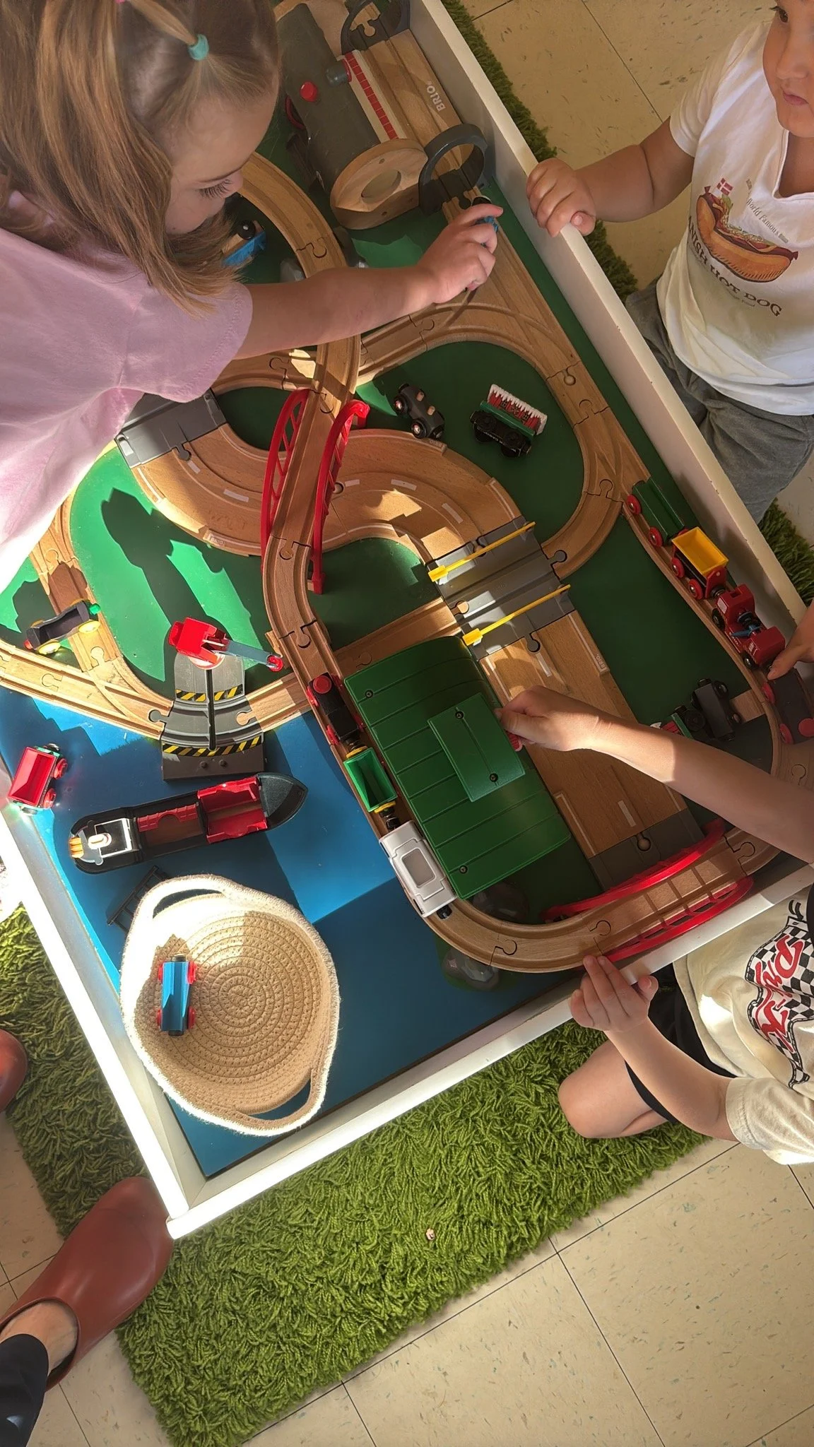 Children playing with a wooden train set on a blue and green table, with some children wearing casual clothes and hats, and a green shaggy rug underneath the table.