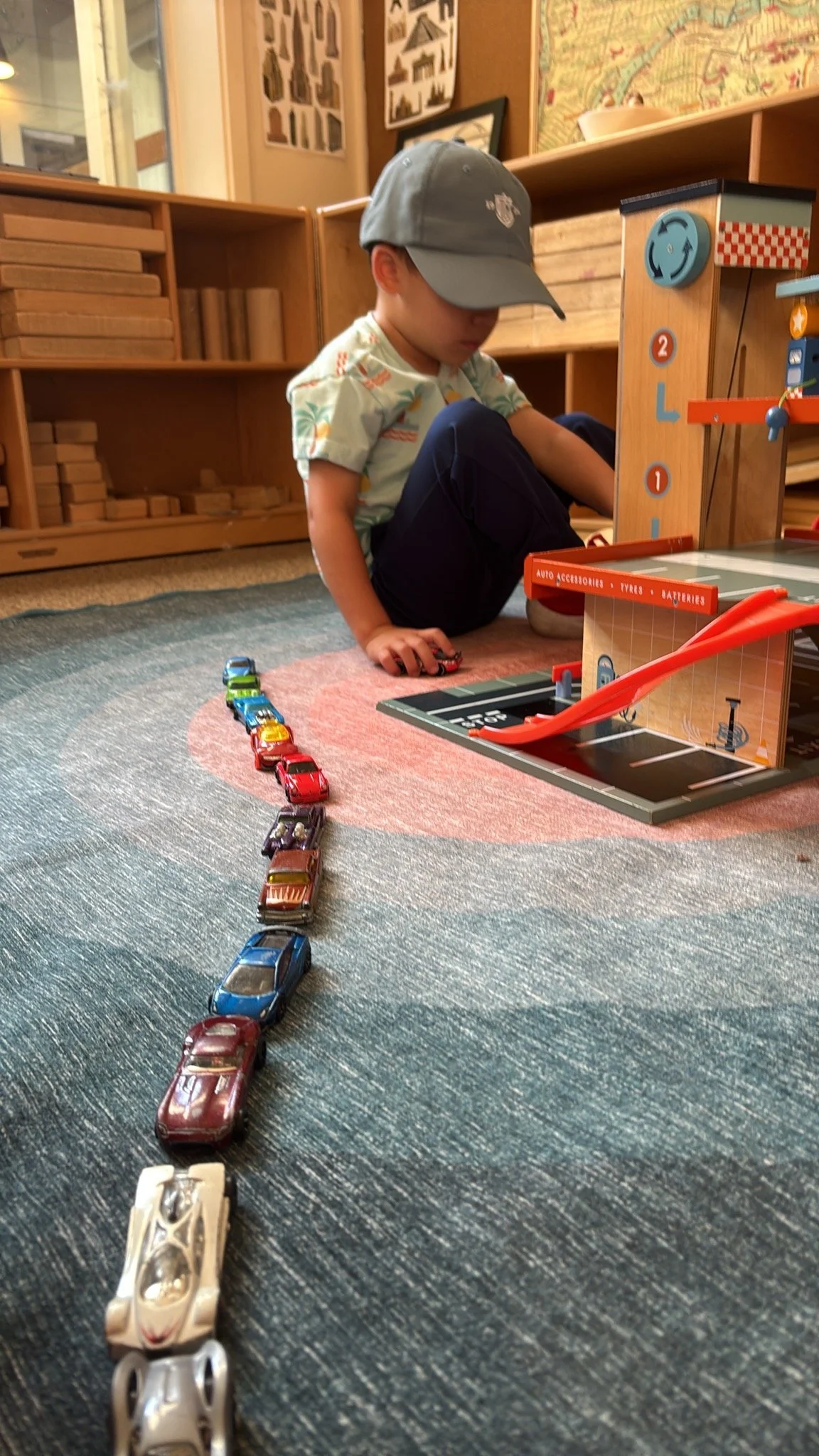 A young boy wearing a gray cap and a colorful shirt is sitting on a carpeted floor playing with toy cars. There is a line of toy cars in front of him and a wooden parking garage with a ramp and parking spaces nearby. Wooden shelves with storage boxes