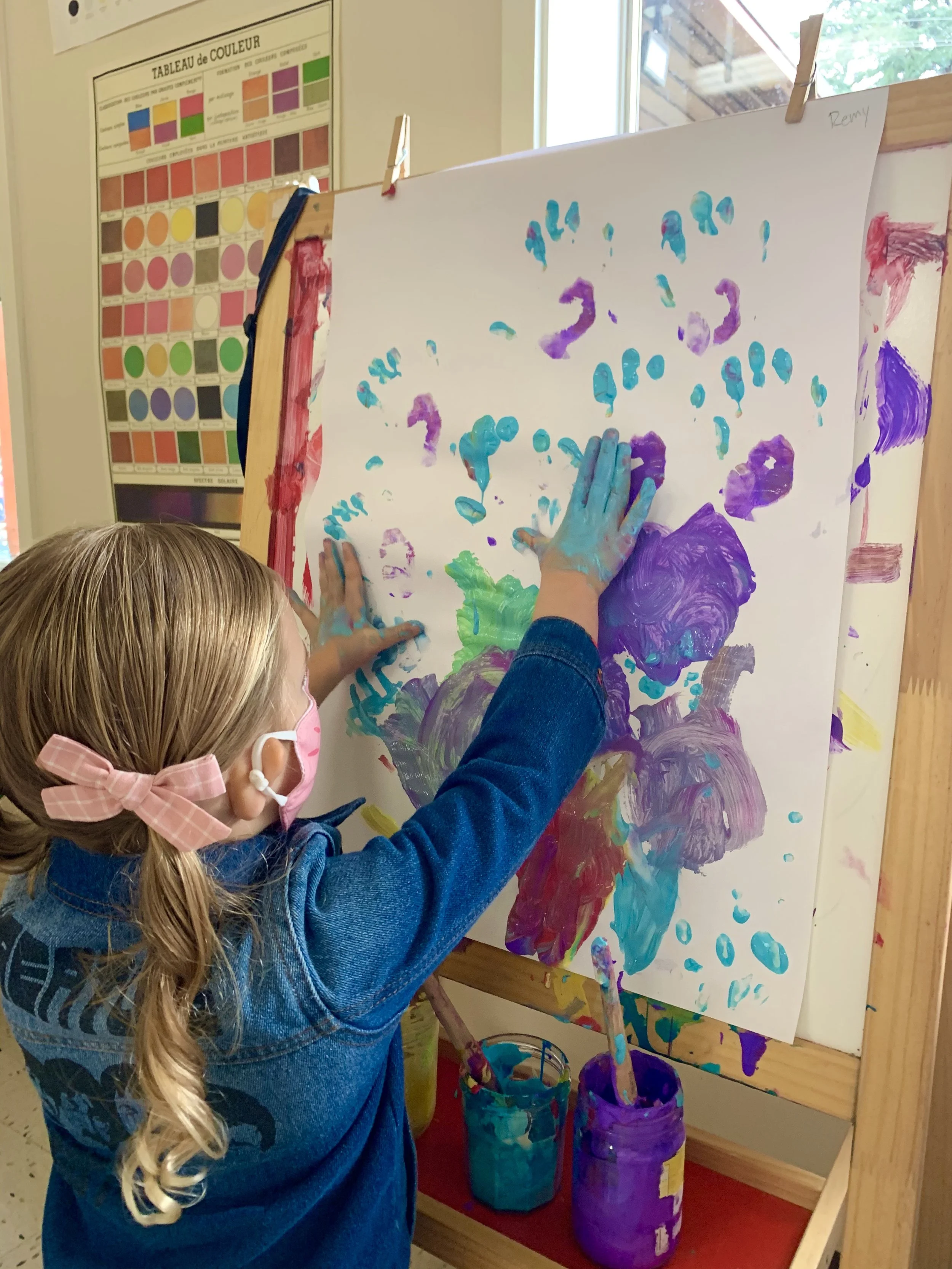 Child with long blonde hair and pink mask pressing hands onto a colorful abstract painting on an easel in a classroom, with paint buckets underneath, a color chart on the wall, and a window showing trees outside.