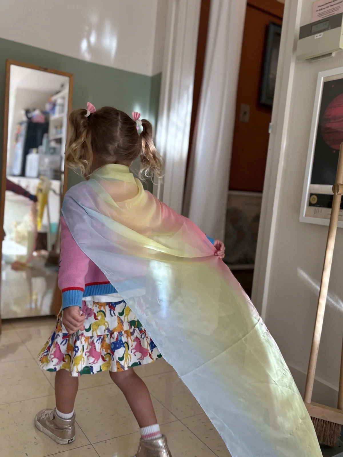 A young girl with pigtails wearing a colorful unicorn print skirt, a blue and pink sweater, and gold sneakers, holds a large, iridescent rainbow-colored rainbow fabric in a room with a mirror, white walls, and various items in the background.