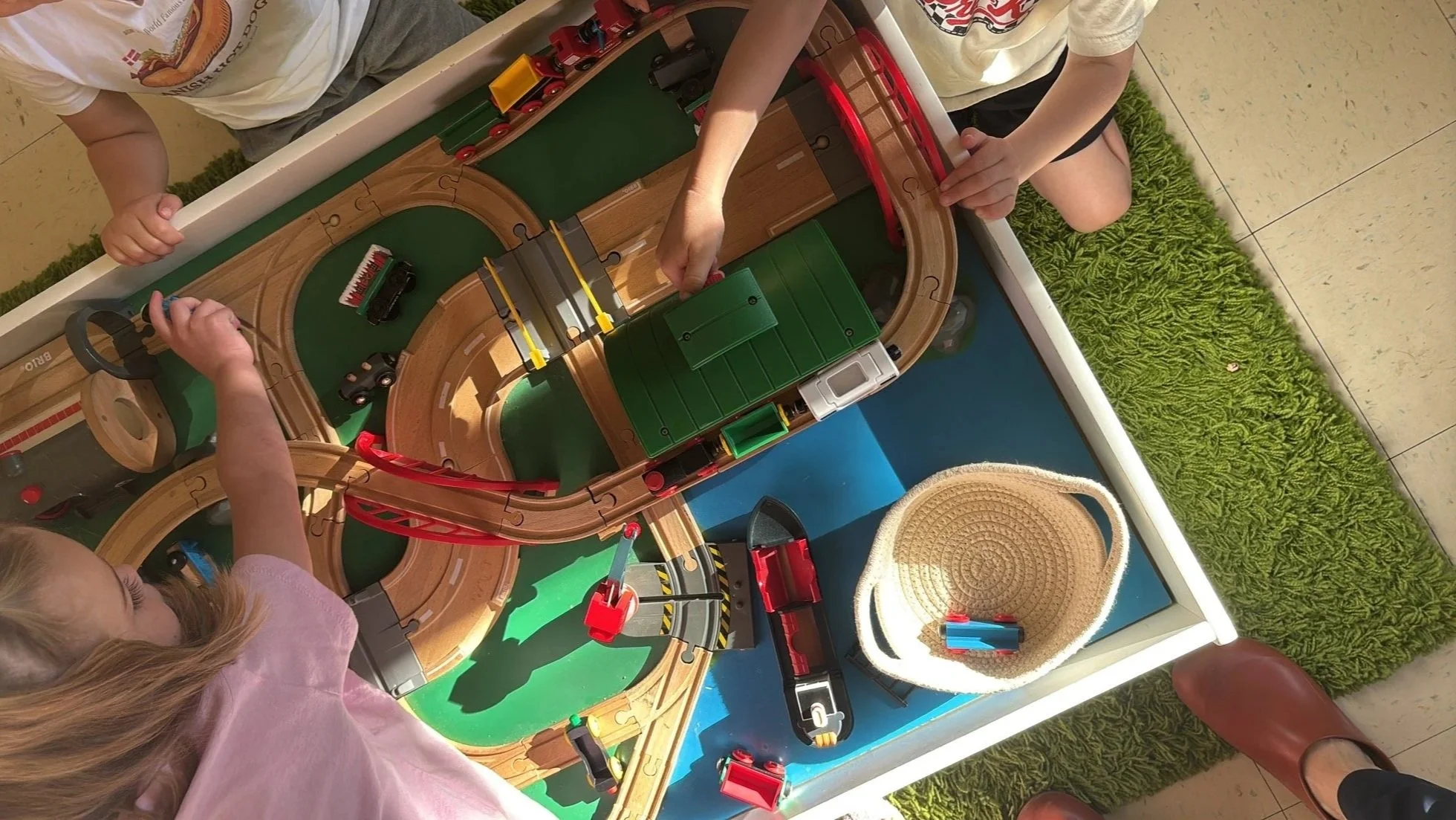 Children playing with a wooden train set inside a white box. The train set includes tracks, trains, and accessories.