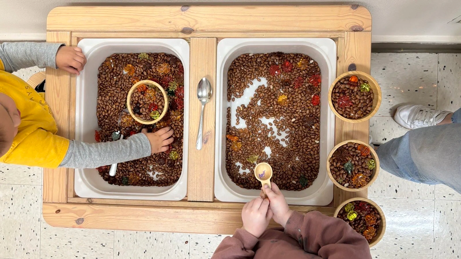 Children playing at a sensory table at Youngset Preschool with dried grains and plastic fall leaves and animals, using small scoops and bowls.