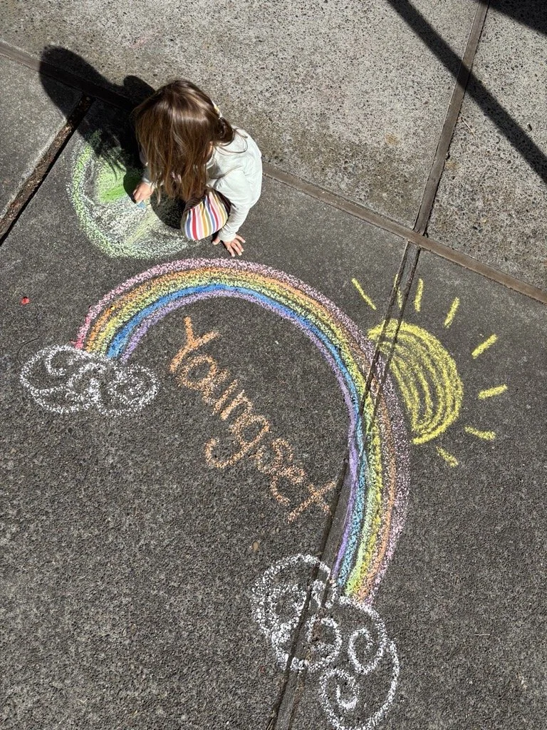 Child drawing a colorful rainbow, sun, clouds, and the words 'Youngset' on the pavement with chalk.